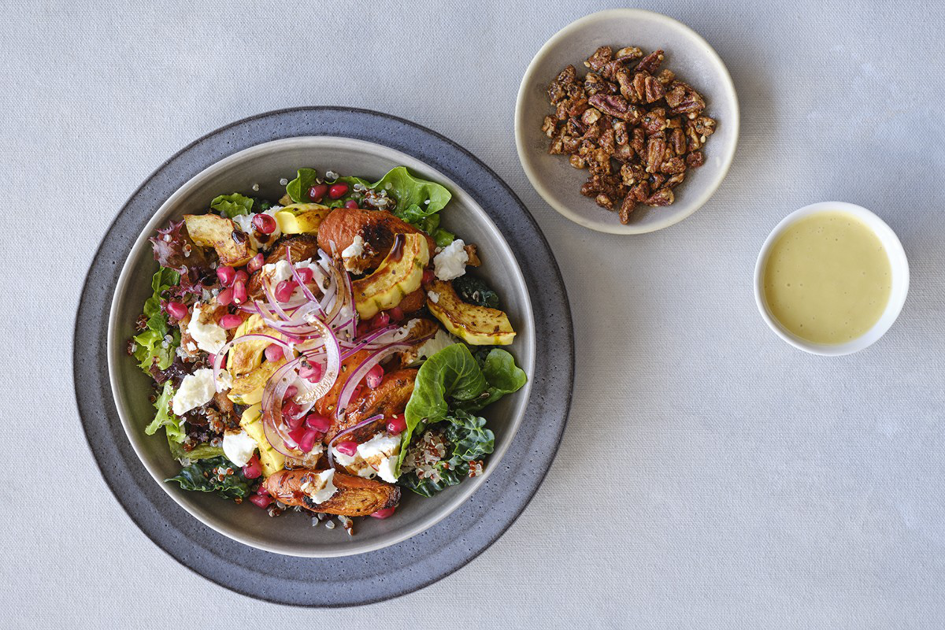 A bowl of mixed greens, roasted squash, pomegranate seeds, goat cheese, and quinoa is served alongside a small bowl of candied pecans and a cup of creamy dressing.