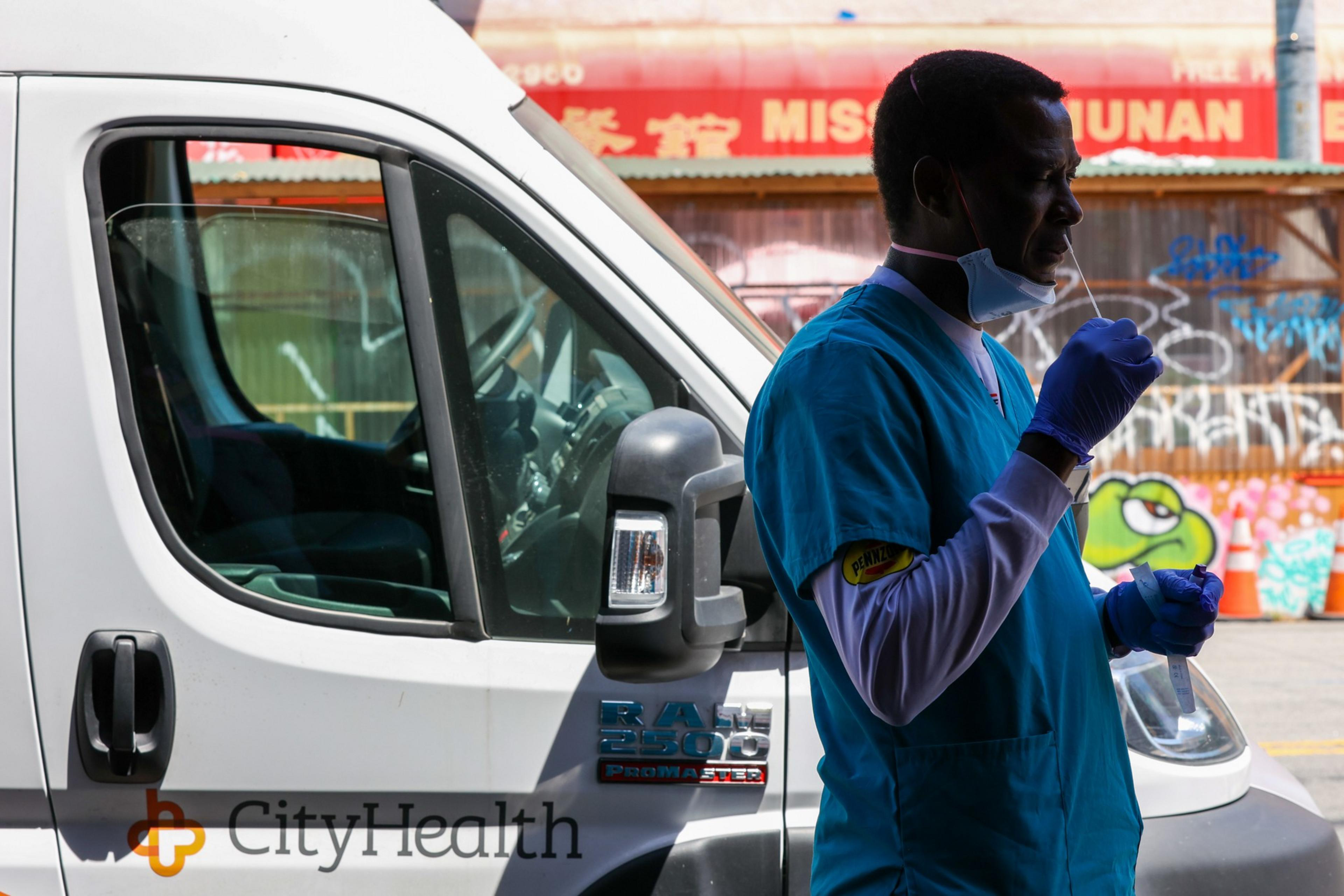 A healthcare worker in blue scrubs and purple gloves prepares to administer a COVID-19 nasal swab test next to a white medical van.