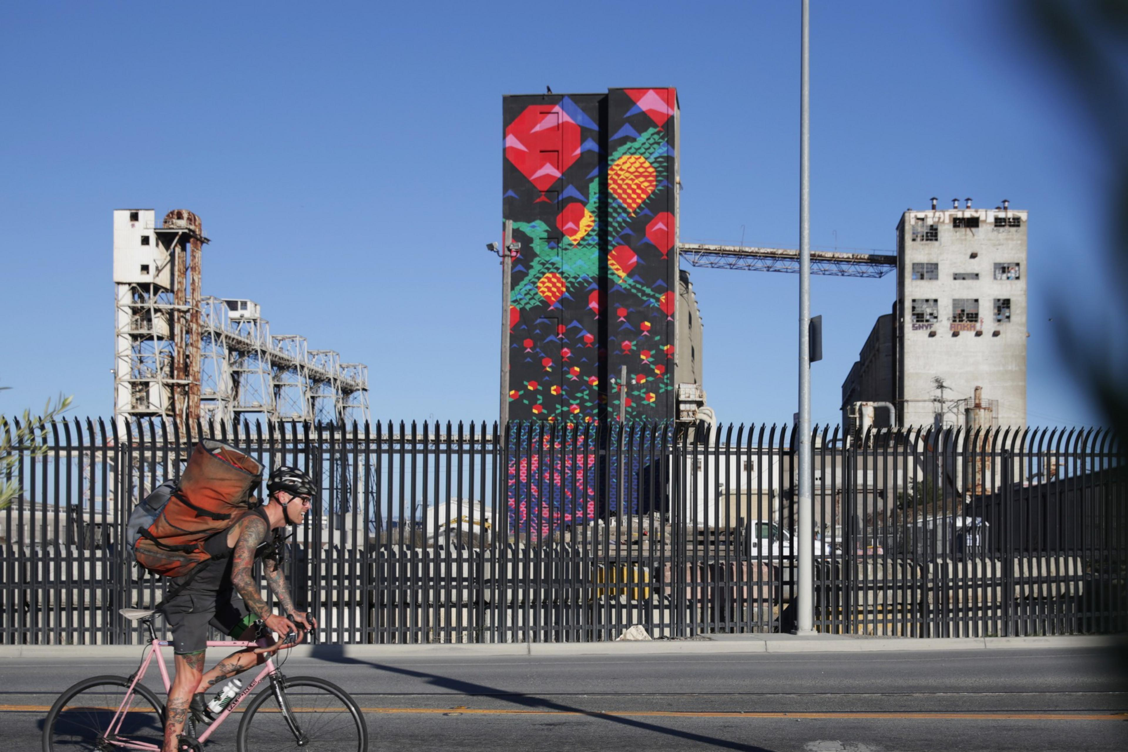 A cyclist with a backpack rides on a street next to a fence. Behind the fence are industrial buildings, including a tall, colorful mural painted with geometric shapes.
