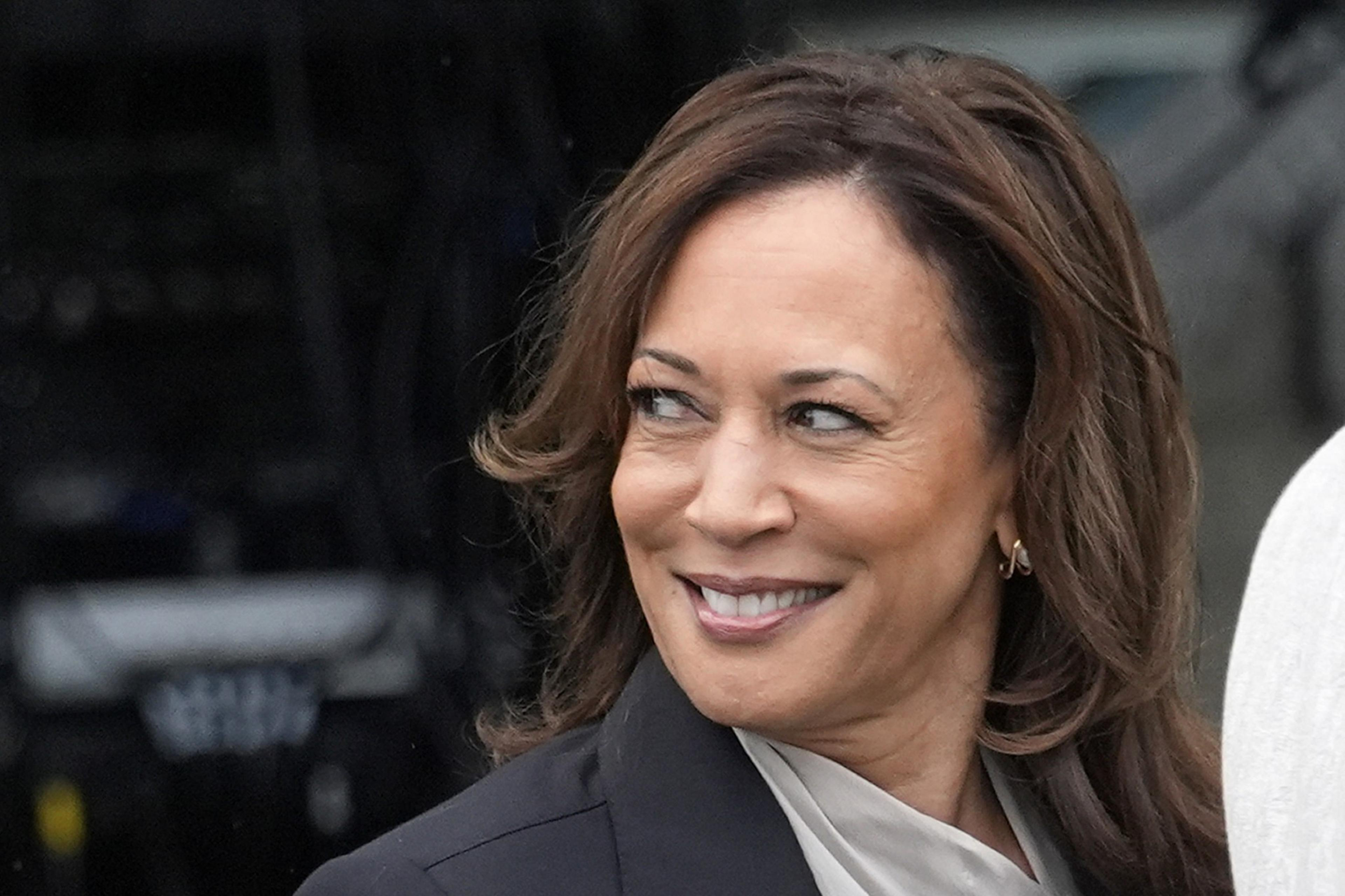 A woman with shoulder-length brown hair smiles while looking to her left. She is wearing a black blazer and has small hoop earrings. The background is indistinct.