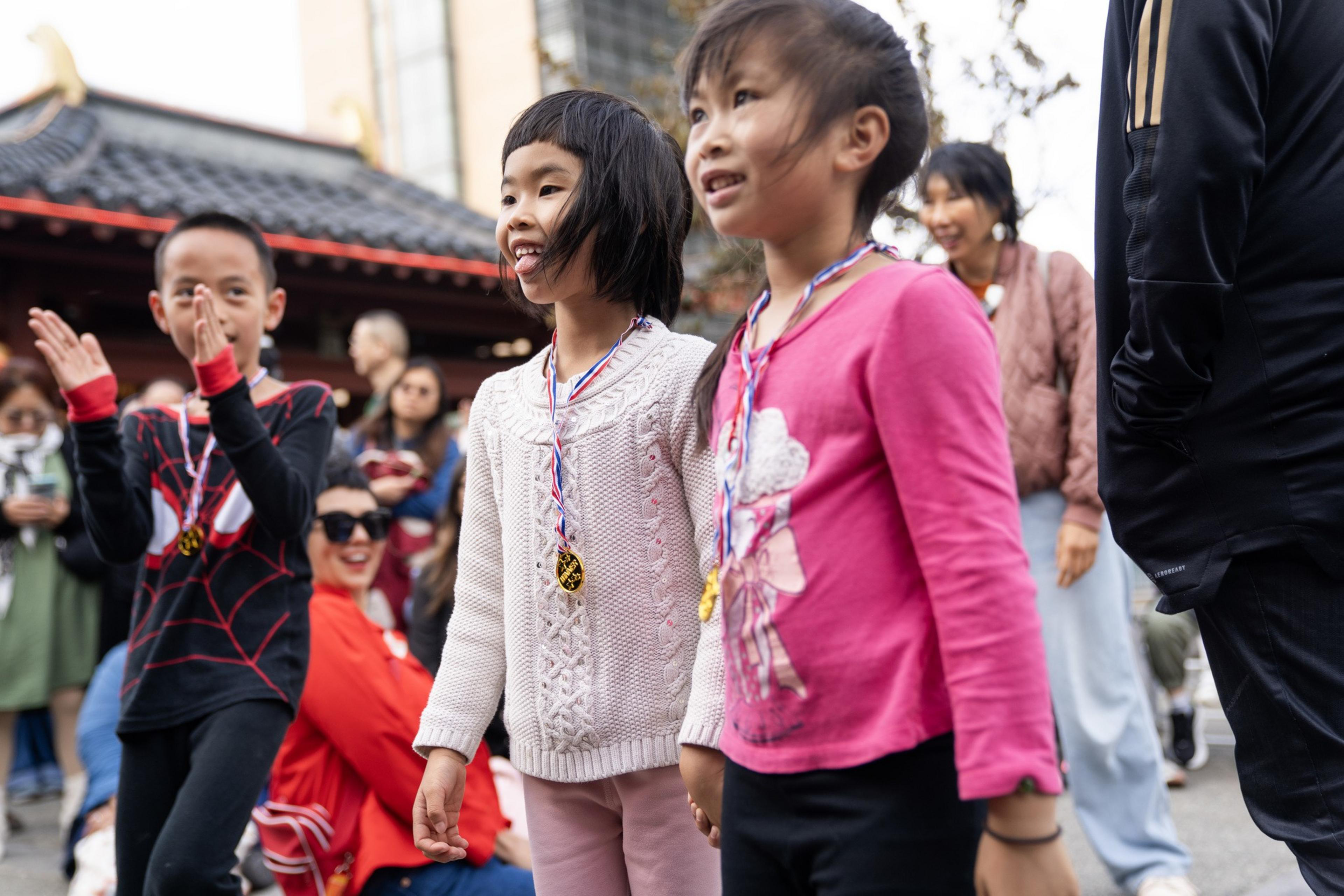 Three children wearing medals stand smiling, with a crowd of people behind them in an outdoor setting.