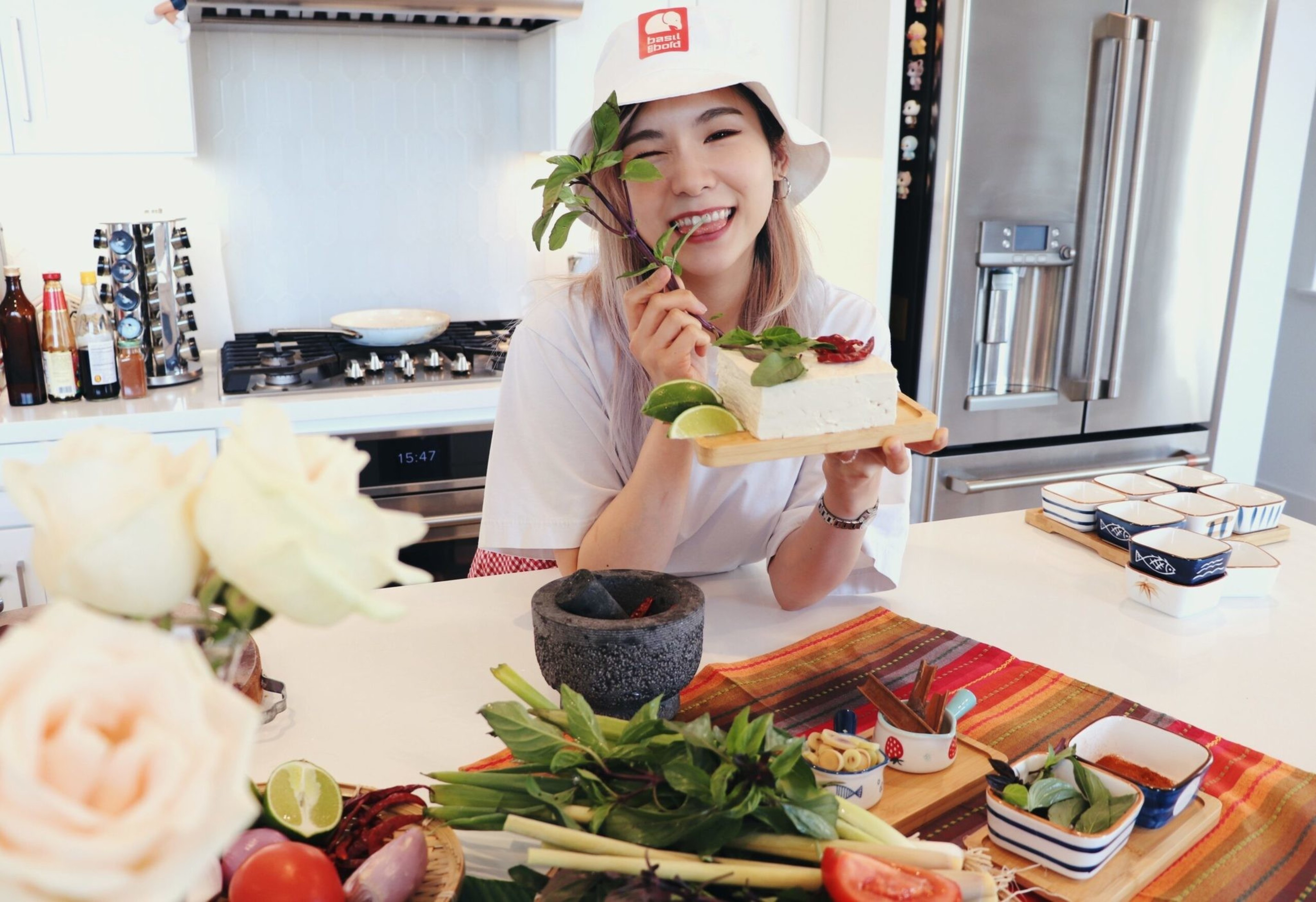 A smiling woman in a white hat holds a wooden board with tofu, lime slices, and chili, surrounded by fresh herbs and vegetables on a kitchen counter.