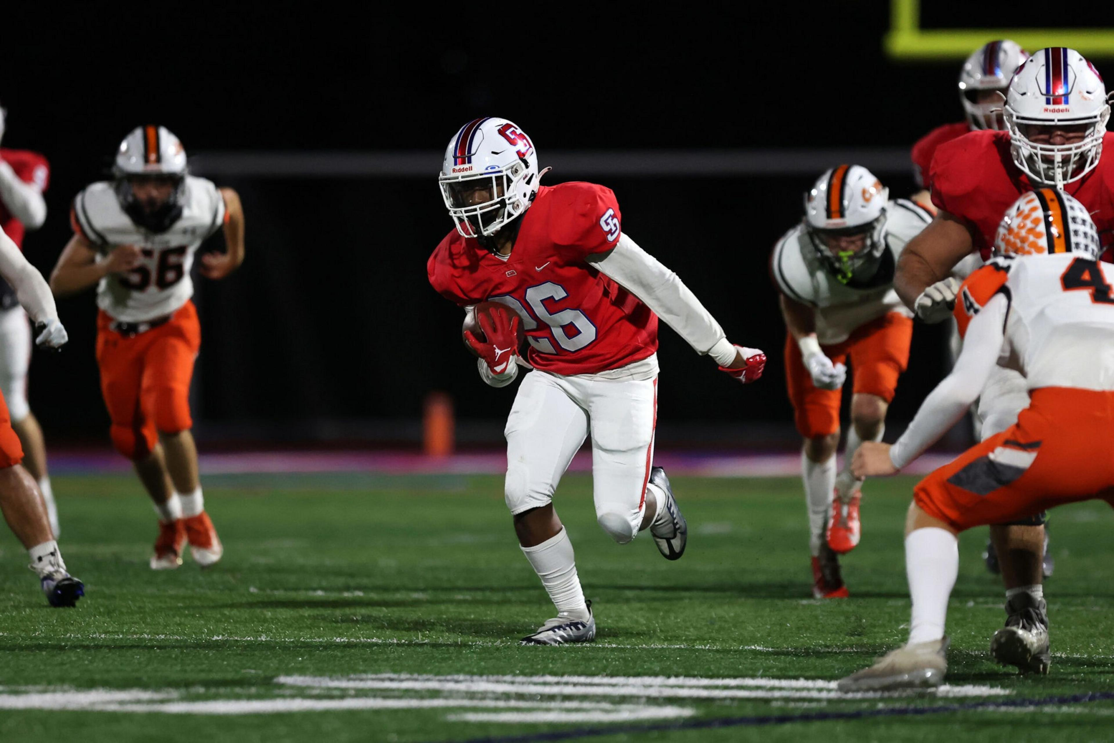 A football player in a red jersey with number 56 runs with the ball while players in white and orange uniforms chase him on the field.