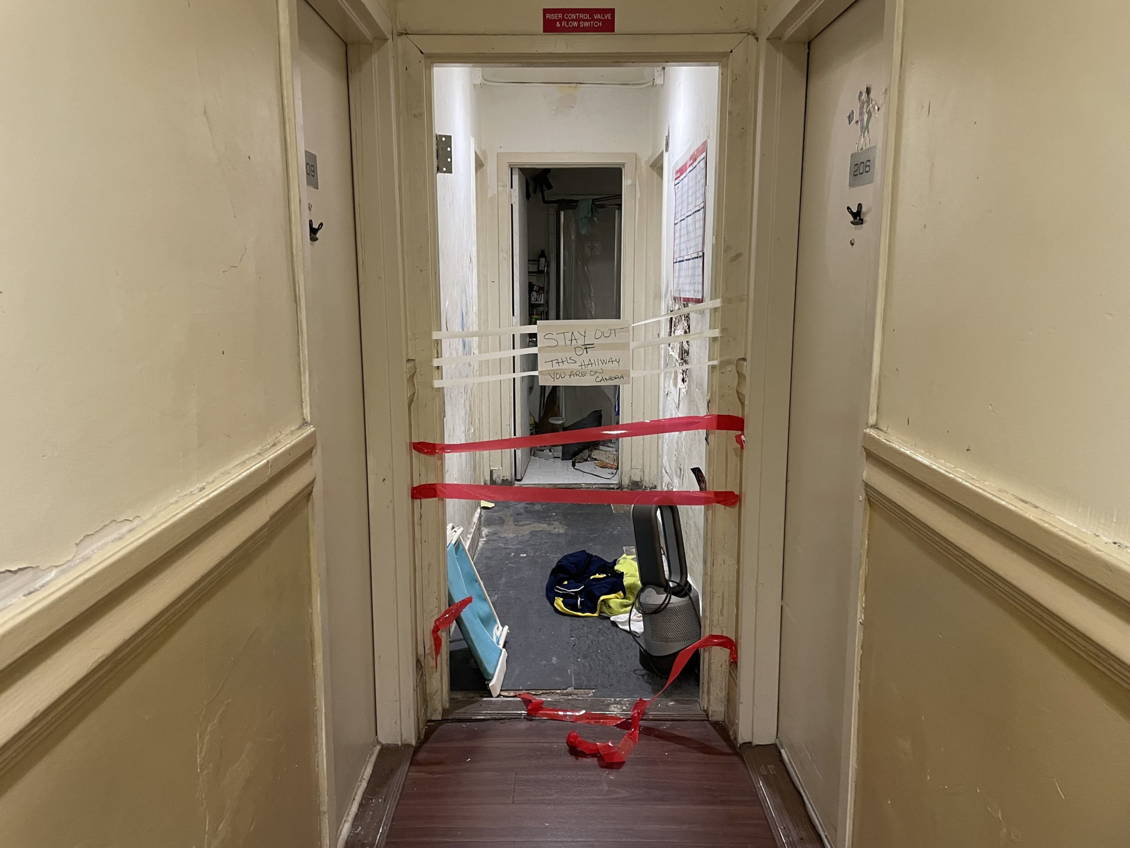 A hallway with two doors is blocked by red tape and a sign saying &quot;Stay out.&quot; Inside, there are various items on the floor, and the area appears under construction.