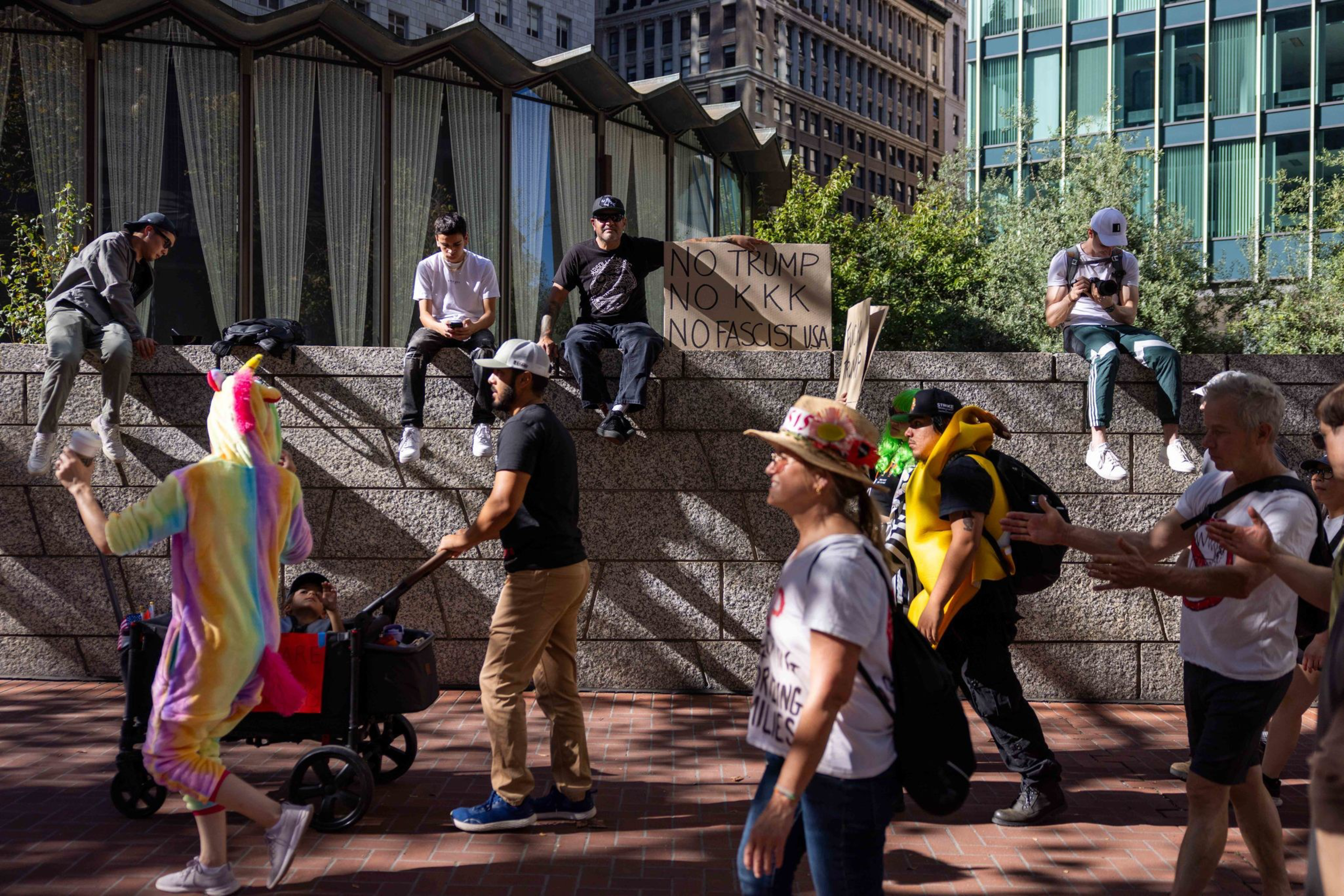 People walk and sit near a stone wall in an urban area, with one holding a sign that reads “No Trump, No KKK, No Fascist USA.”
