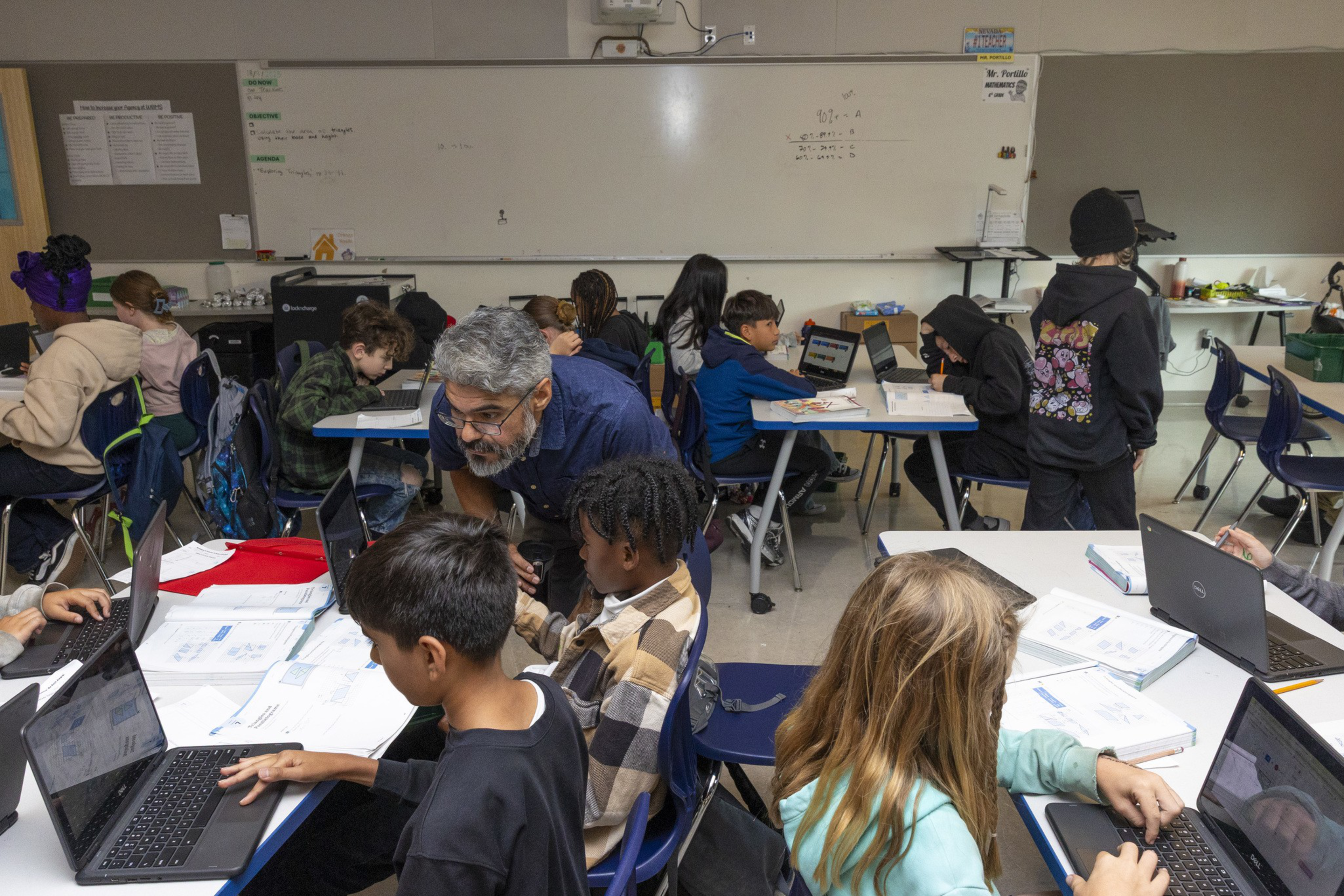 Students work on laptops and notebooks at desks in a classroom while a teacher leans over to assist two students in the foreground.