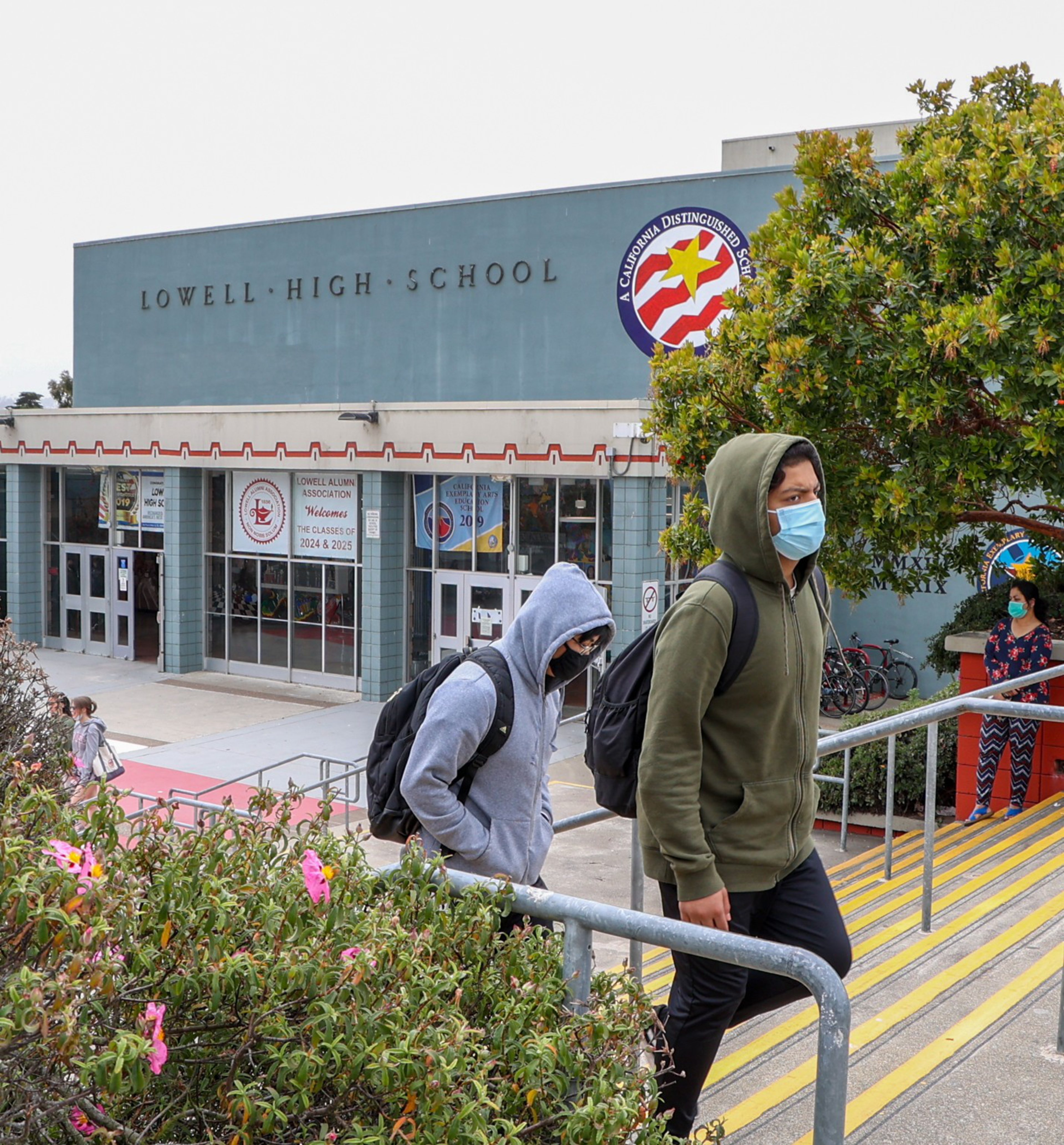 Two students wearing hoodies and face masks walk up stairs outside Lowell High School, which has a sign celebrating it as a California Distinguished School.