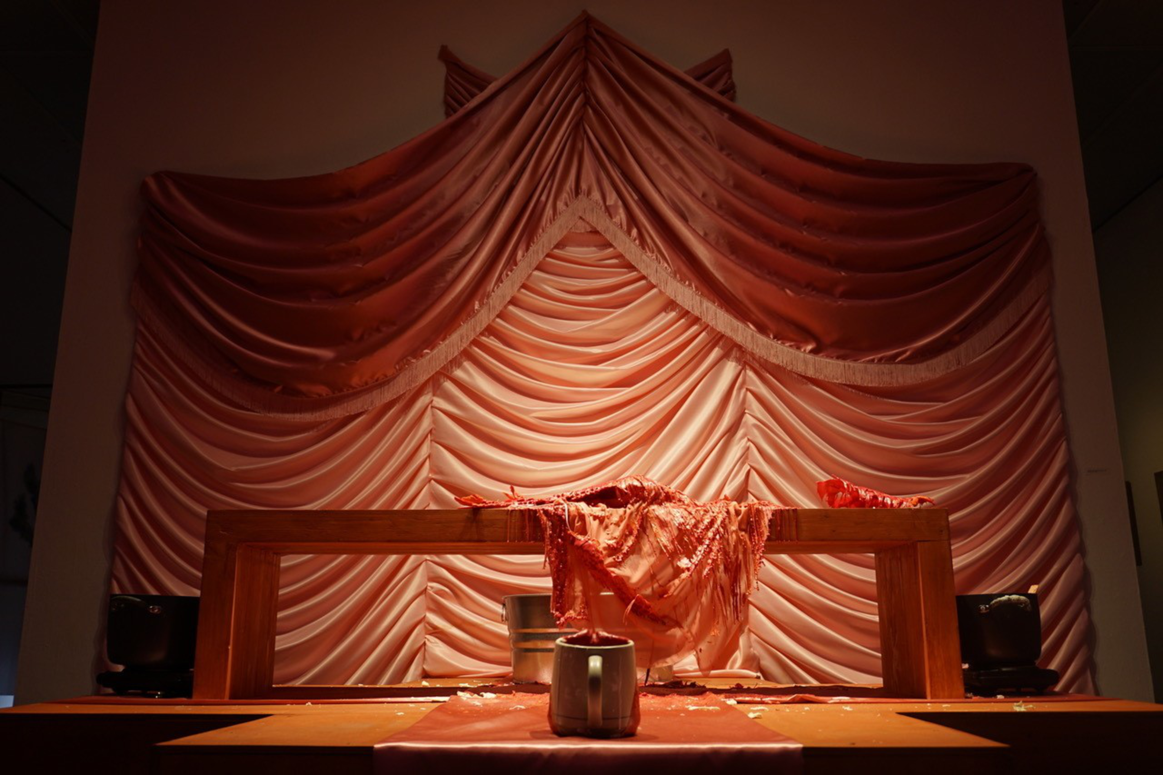 A wooden frame with a draped red cloth is centered in front of a large layered red satin fabric backdrop, with two dark containers on each side.