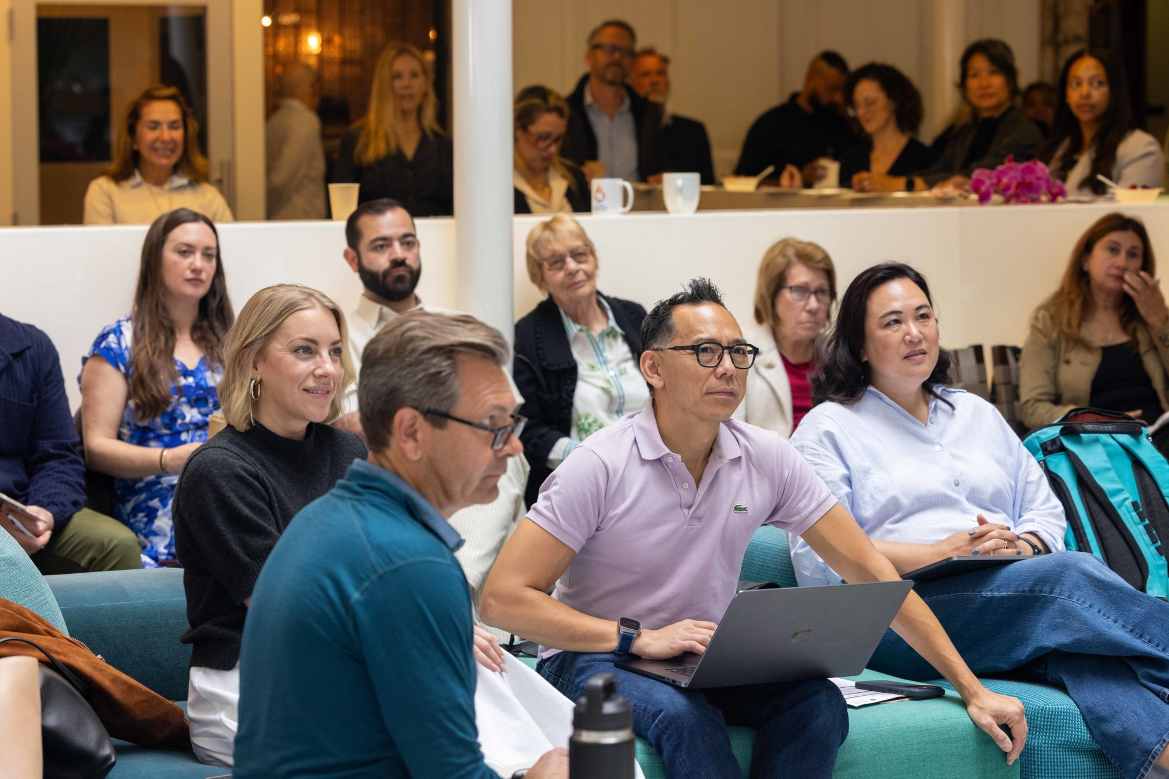 A diverse group of people attentively listens during a meeting, with some taking notes or using laptops in a modern, casual setting.
