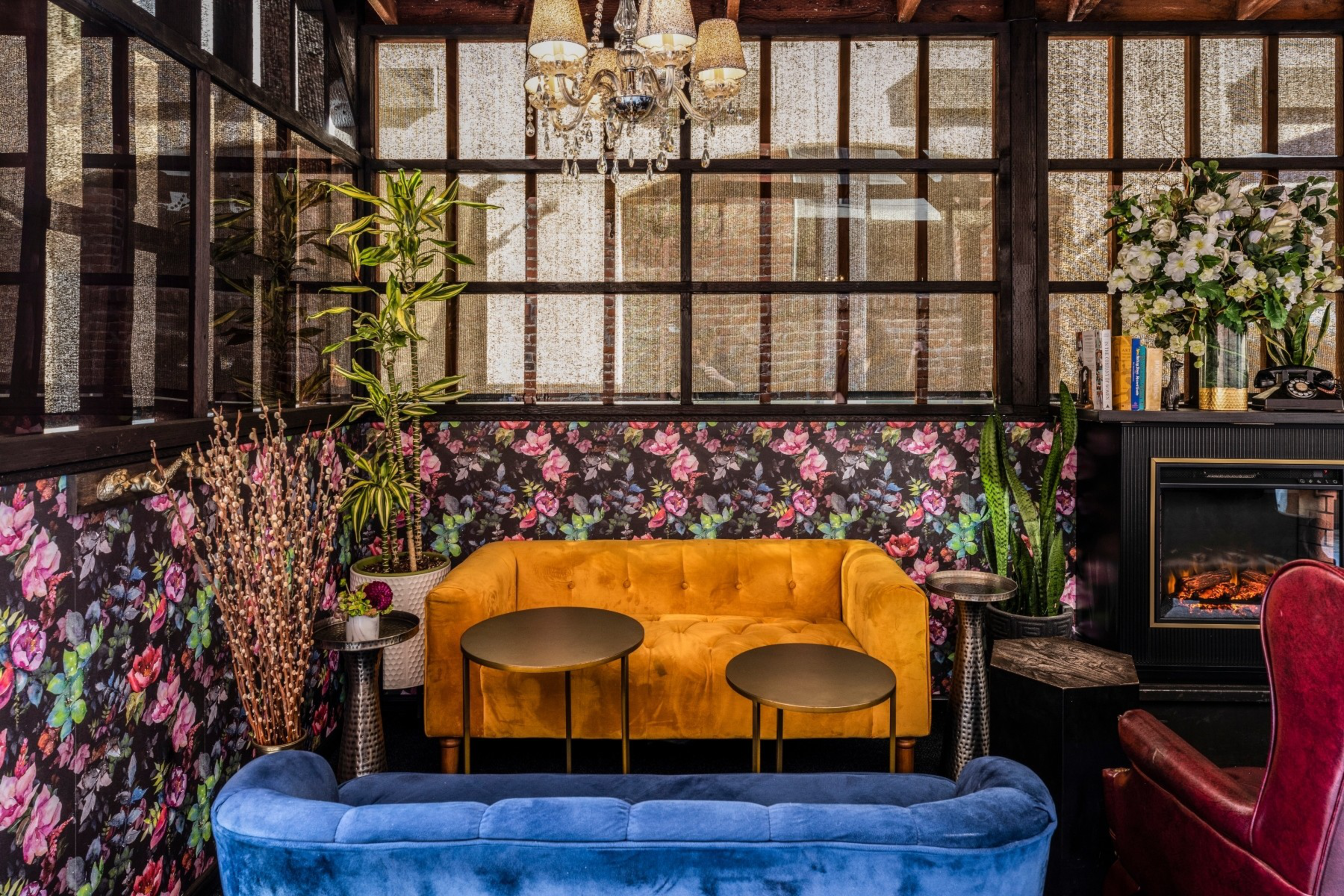 A cozy seating area with a mustard yellow sofa, blue velvet couch, floral wallpaper, indoor plants, a chandelier, and a fireplace with books and flowers.