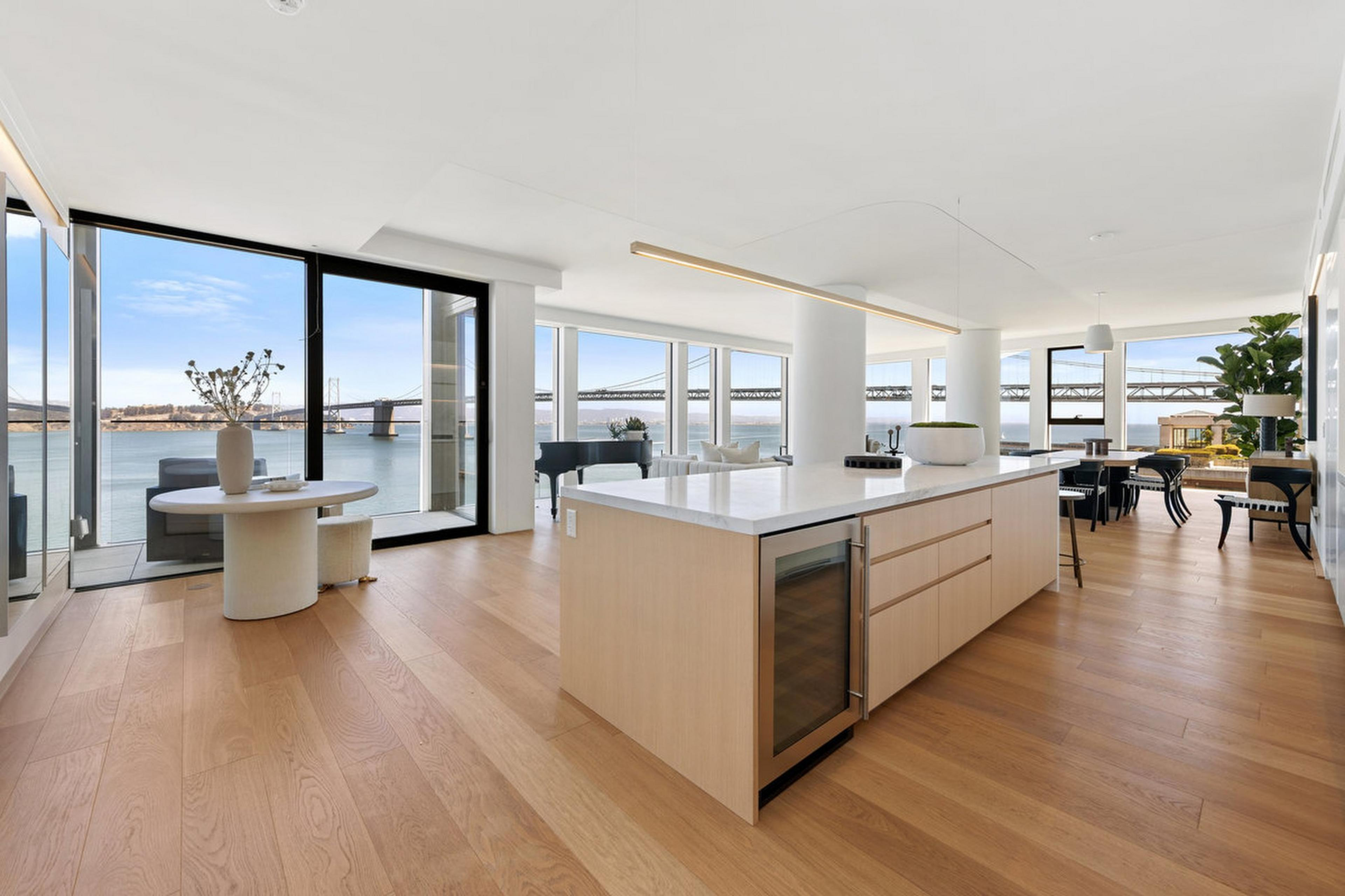 A spacious open kitchen and living area with light wood floors, large island, floor-to-ceiling windows, minimalist furniture, and a waterfront bridge view.