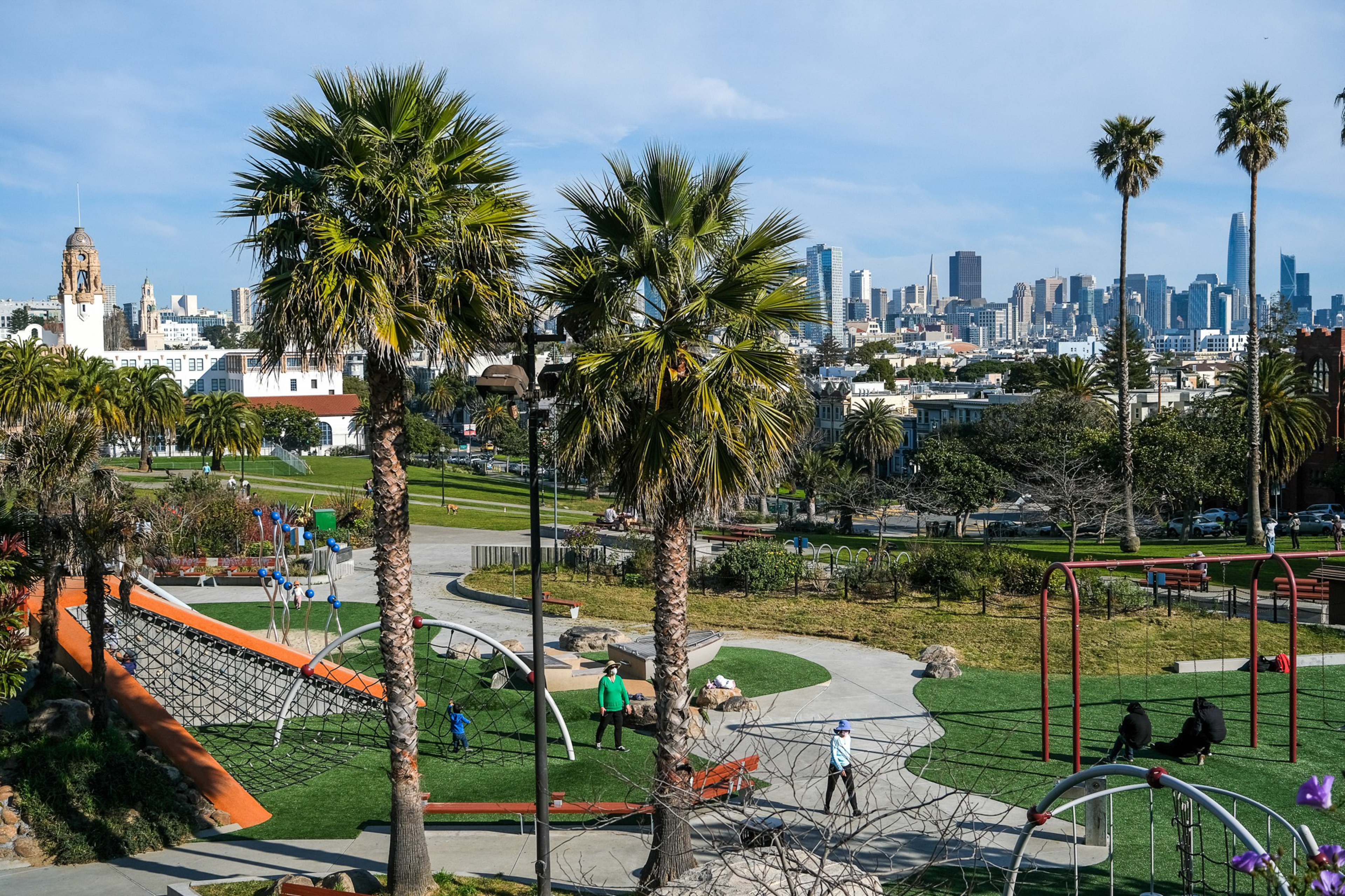 The image shows a vibrant city park with green lawns, palm trees, playground equipment, and people relaxing and playing. In the background, a city skyline rises under a blue sky.
