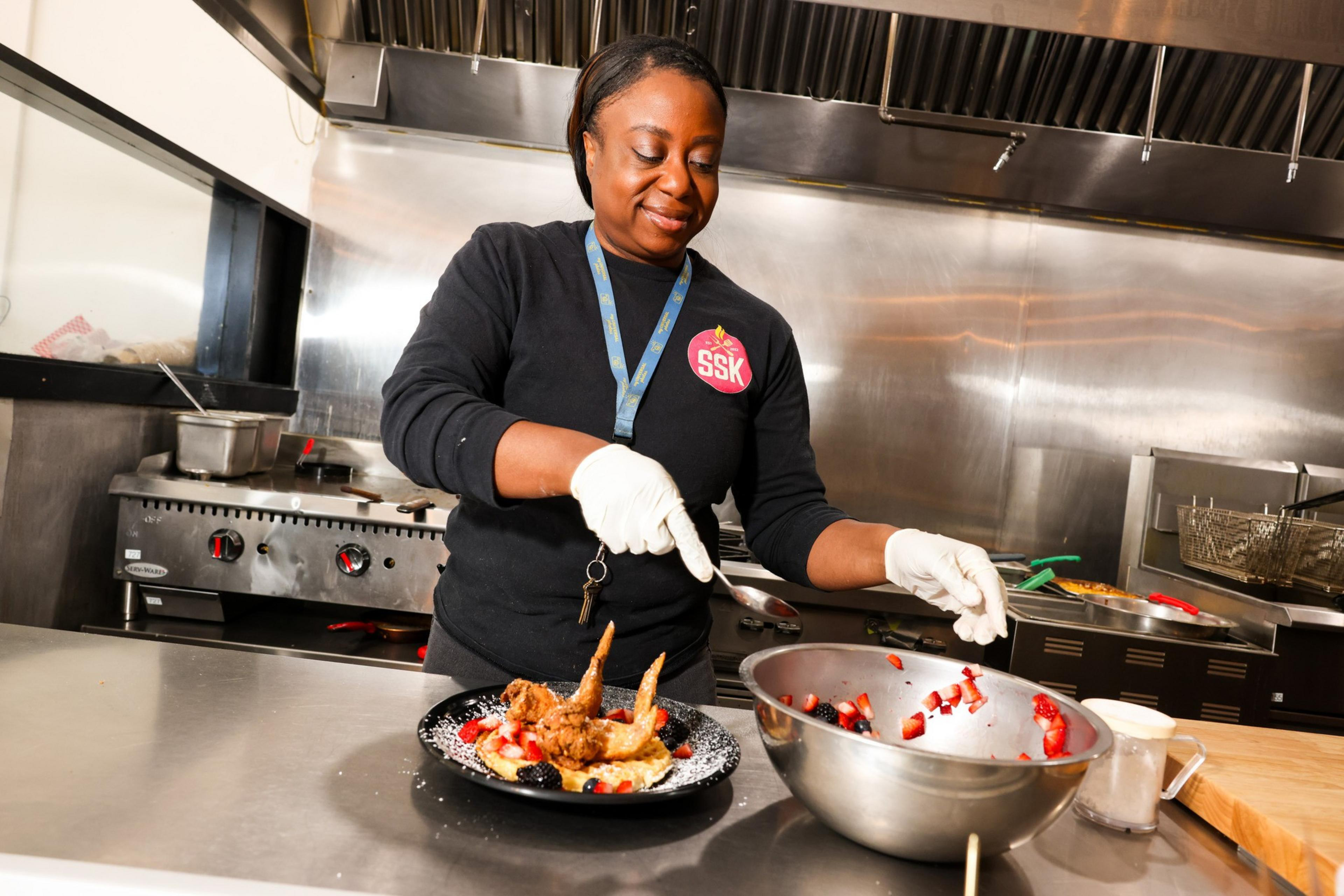 A person wearing gloves is preparing a dish in a kitchen, placing toppings on waffles with fried chicken and berries, with a large bowl of fruit nearby.