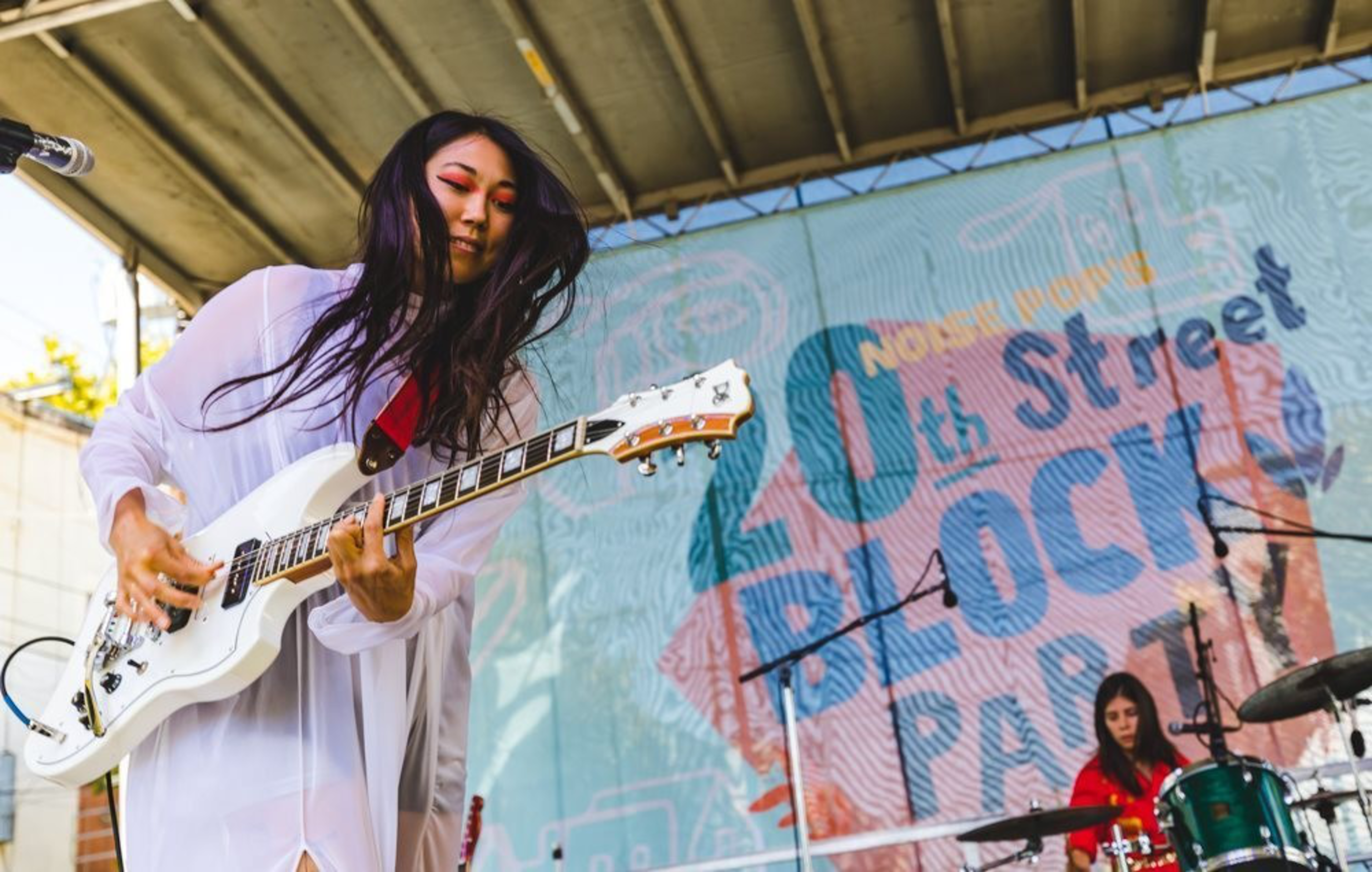 A woman in a white outfit plays a white electric guitar on stage while another person in red plays drums behind her at the 20th Street Block Party.