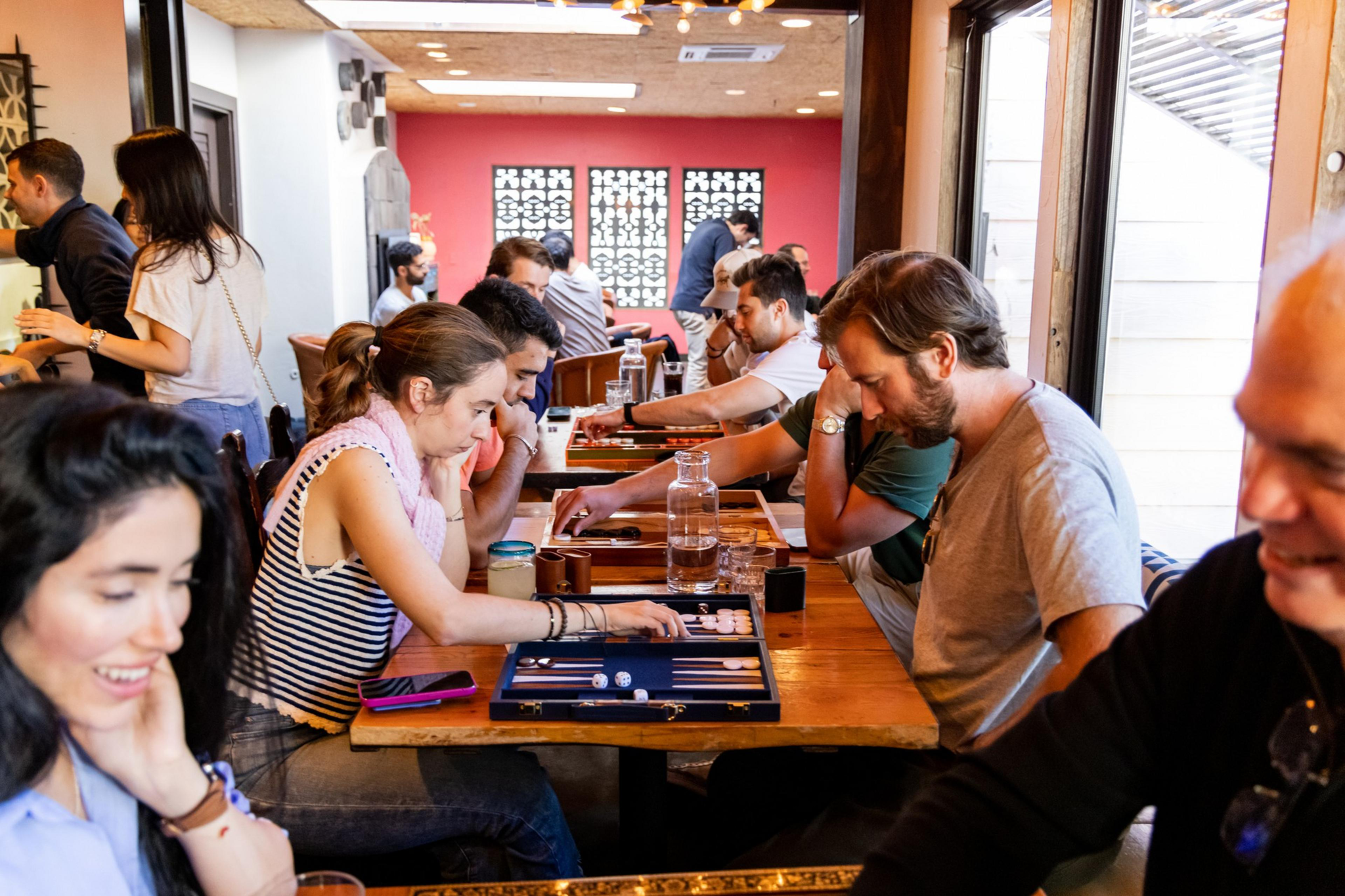People sit closely at long wooden tables playing backgammon, with drinks and personal items scattered around in a bright room with large windows.