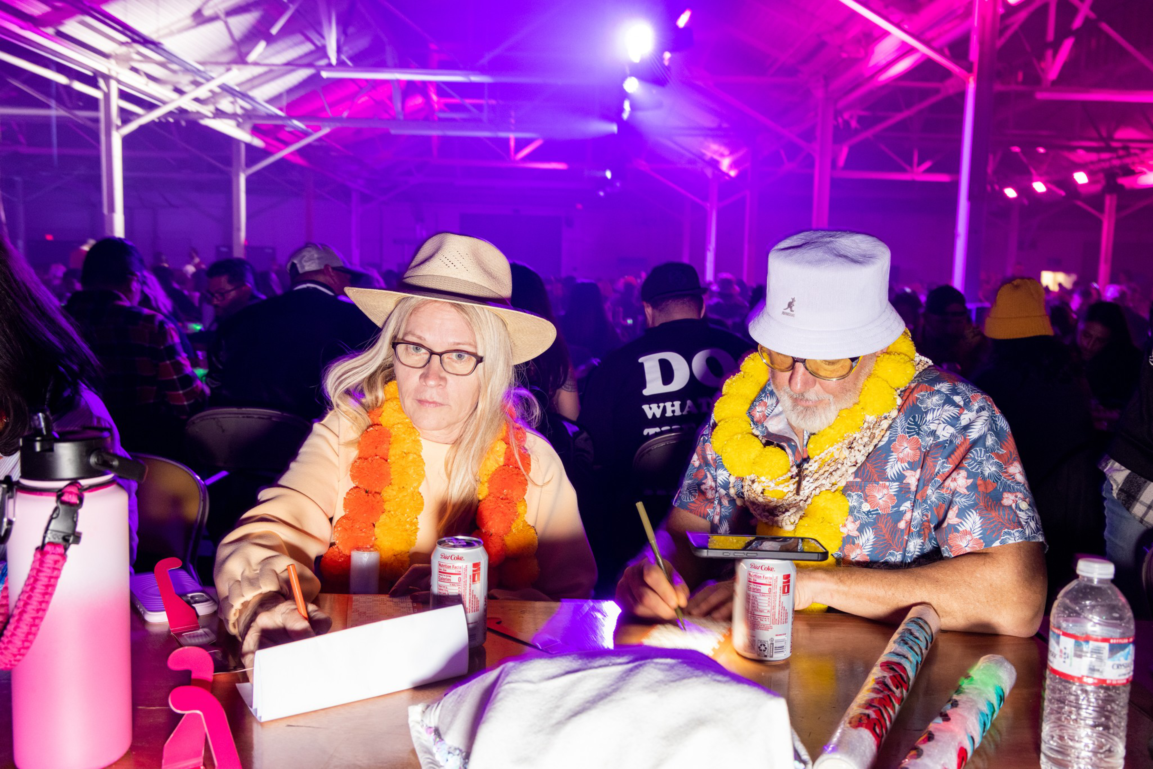 Two people wearing leis and hats sit at a table writing, surrounded by drinks and wrapping paper, with a colorful, crowded background.
