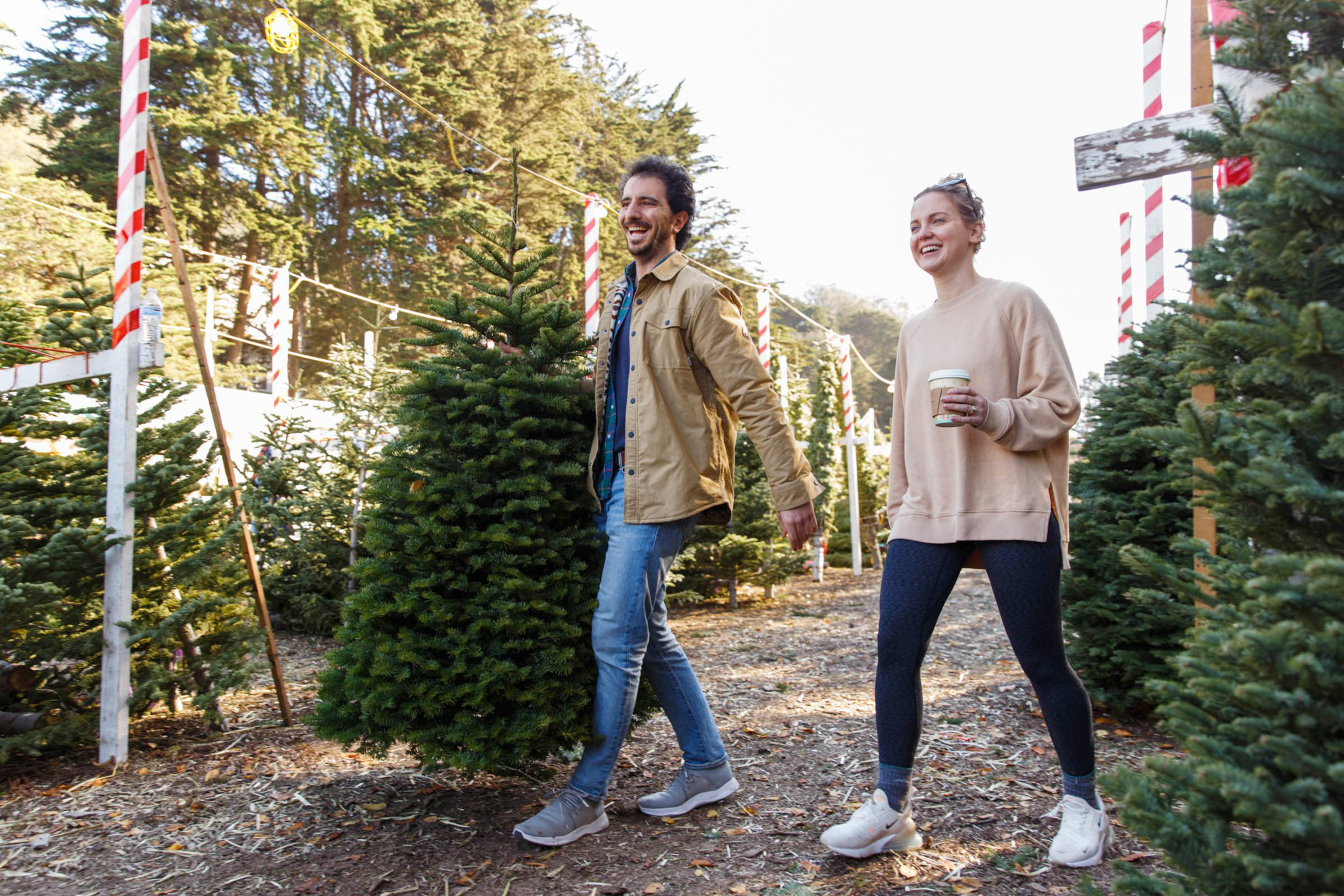 A man and woman walk through a Christmas tree farm, smiling, with the man carrying a freshly cut tree and the woman holding a coffee cup.