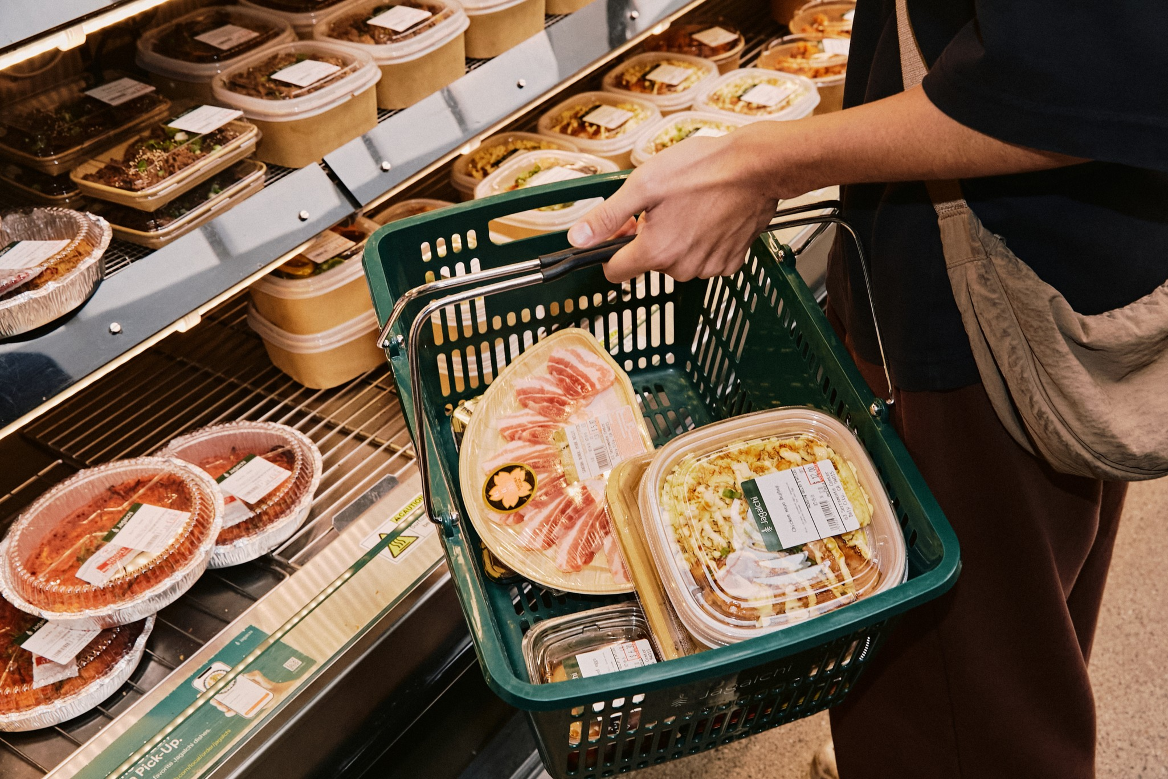 A person holds a green shopping basket containing packaged food, standing next to a refrigerated shelf filled with more packaged meals.