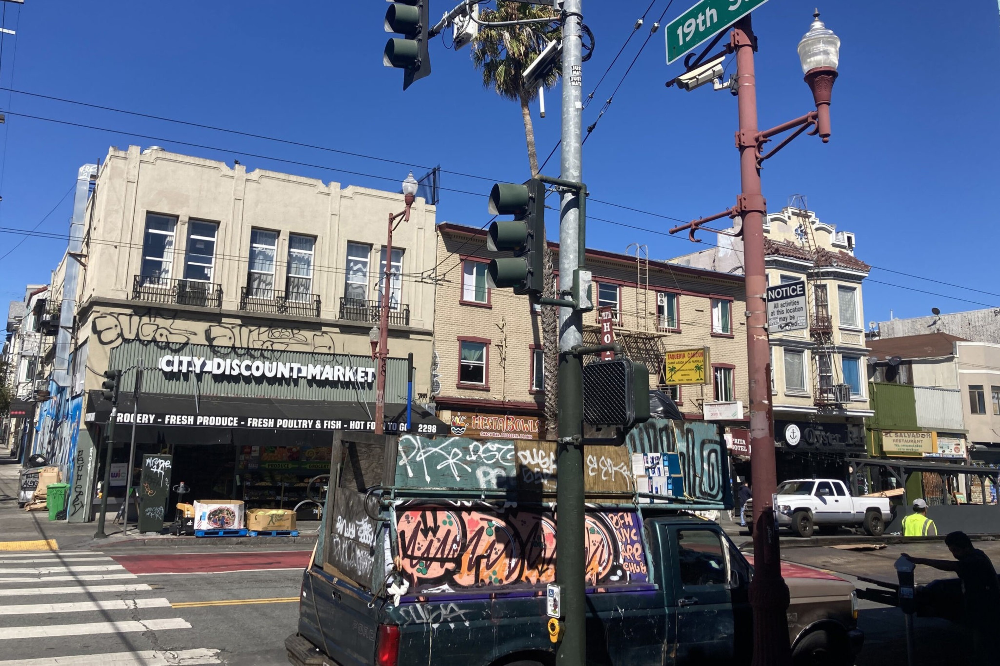 A street corner with a graffiti-covered van, a "City Discount Market" store, various buildings, a palm tree, traffic lights, and a "19th St" sign.