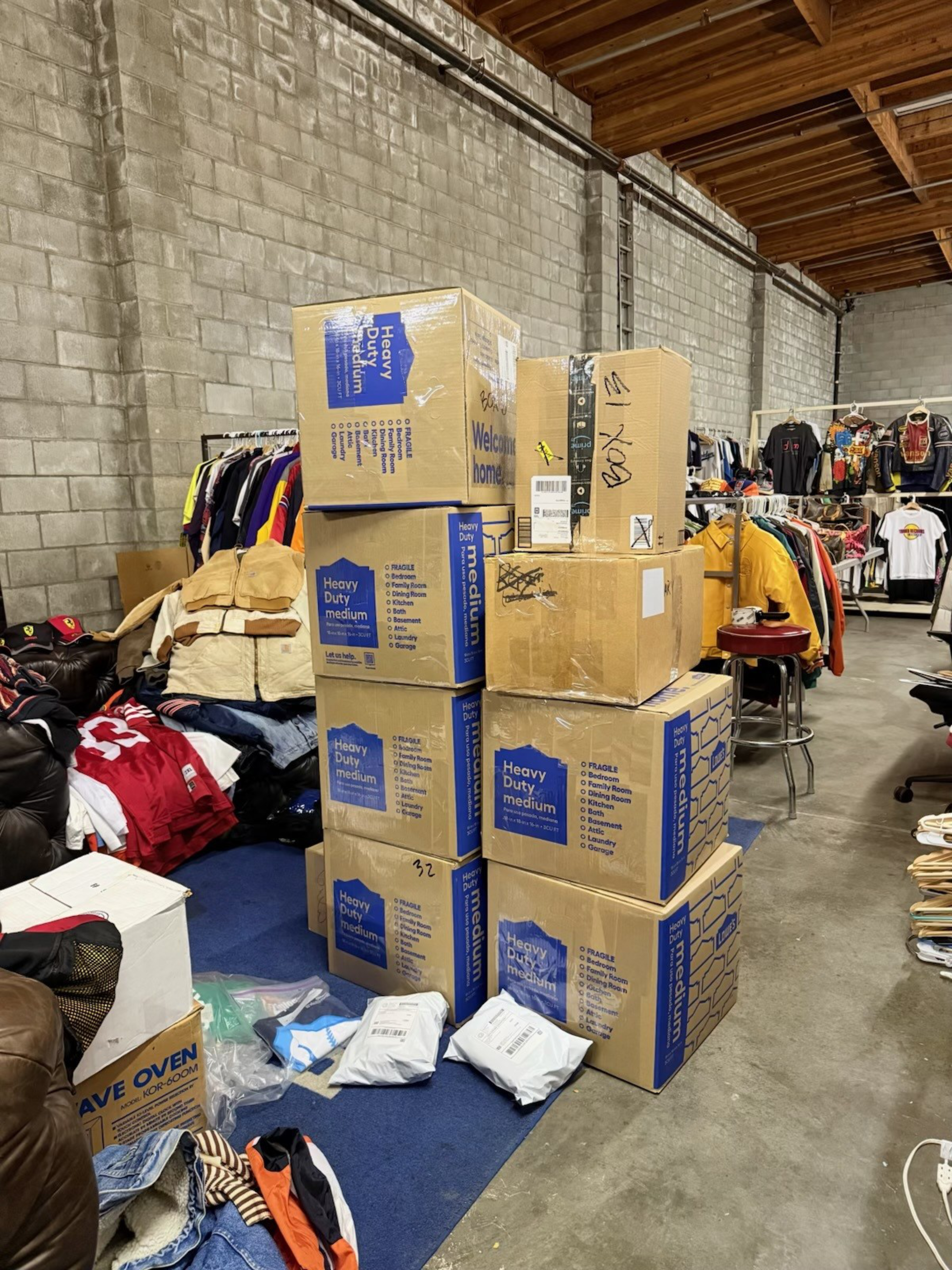 The image shows a stack of heavy-duty cardboard boxes in a warehouse-like space, surrounded by racks of clothing and other scattered packages on the floor.