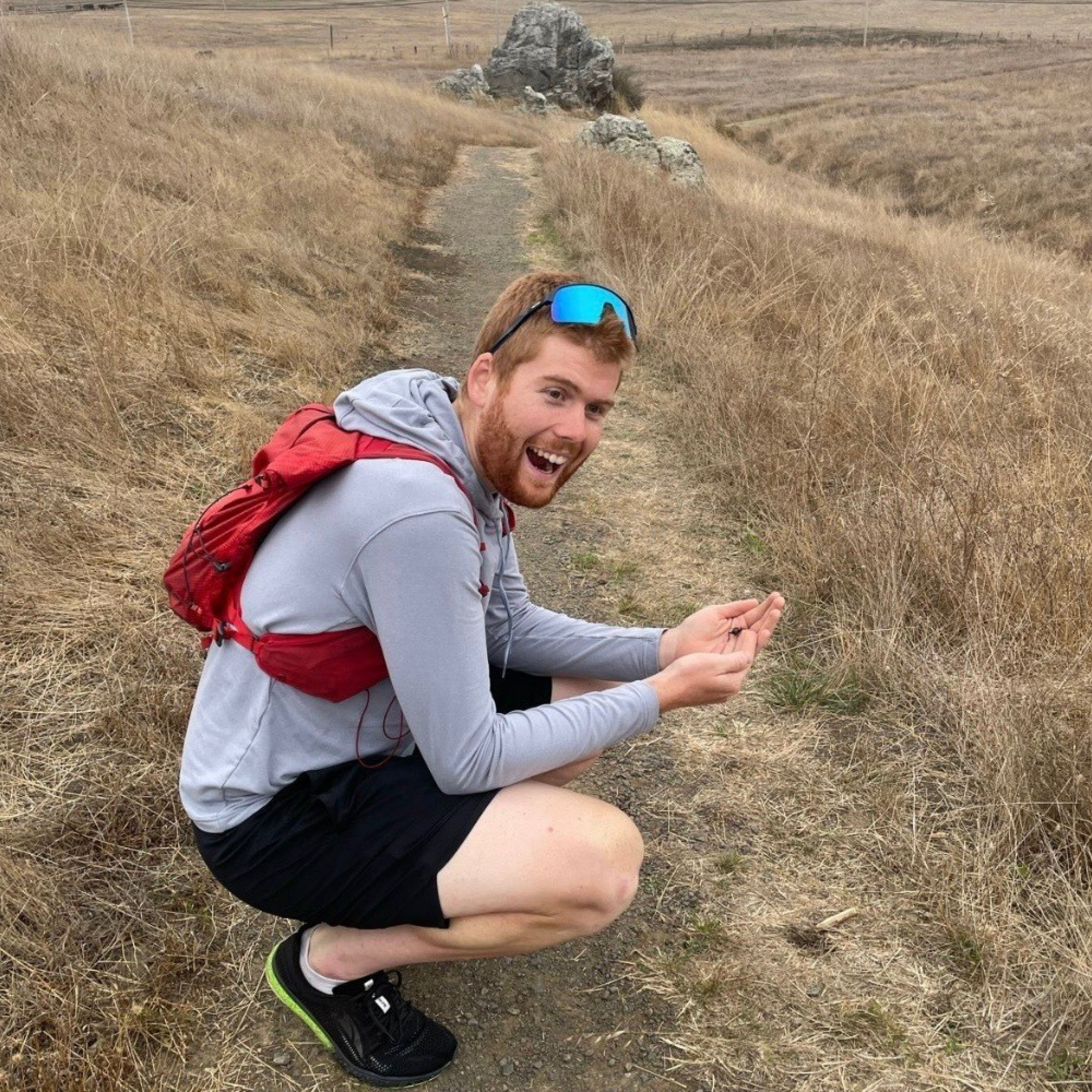 A man wearing a gray hoodie, black shorts, and a red backpack squats on a dry dirt path in a grassy, brown field, smiling and holding something small.
