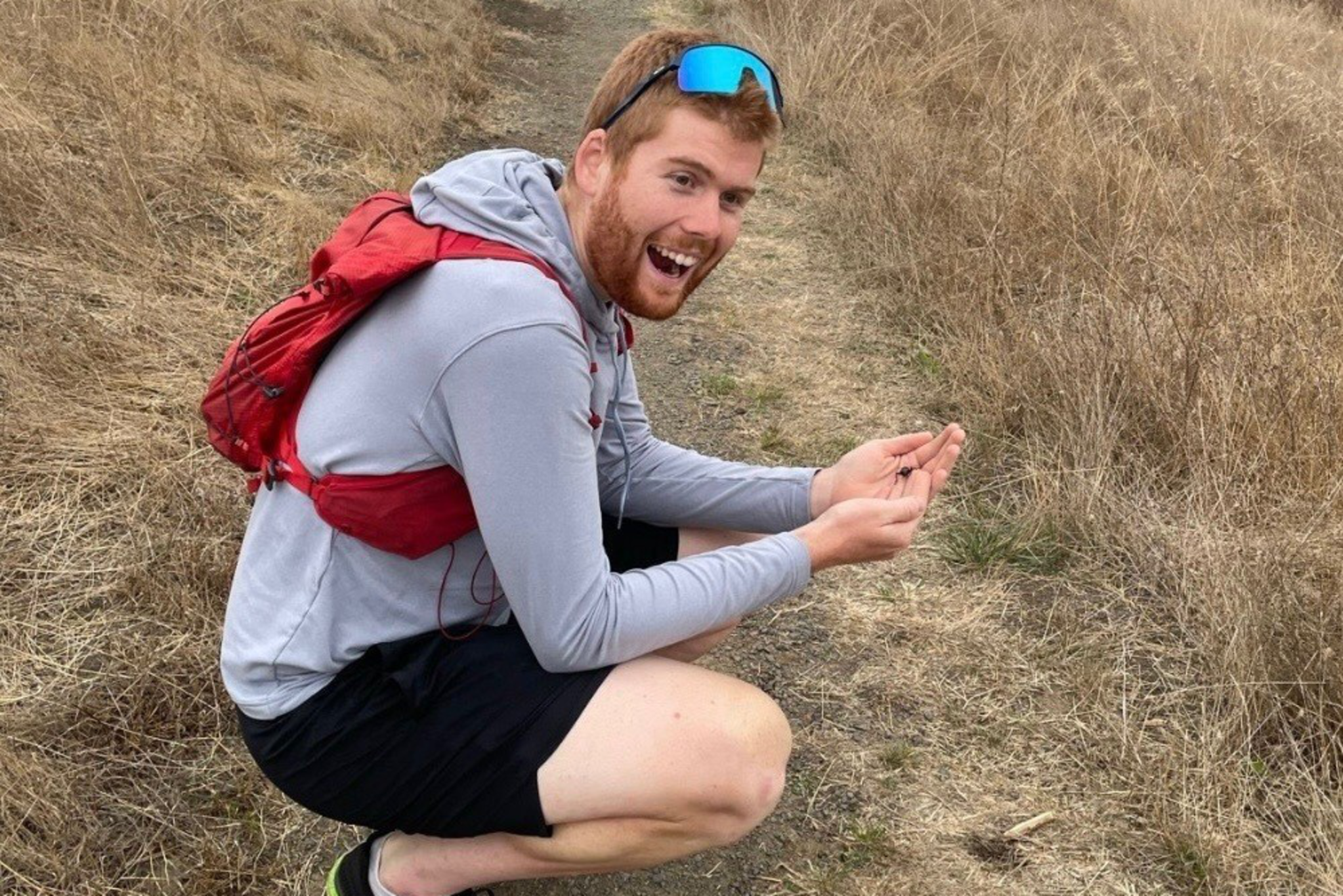 A man wearing a gray hoodie, black shorts, and a red backpack squats on a dry dirt path in a grassy, brown field, smiling and holding something small.