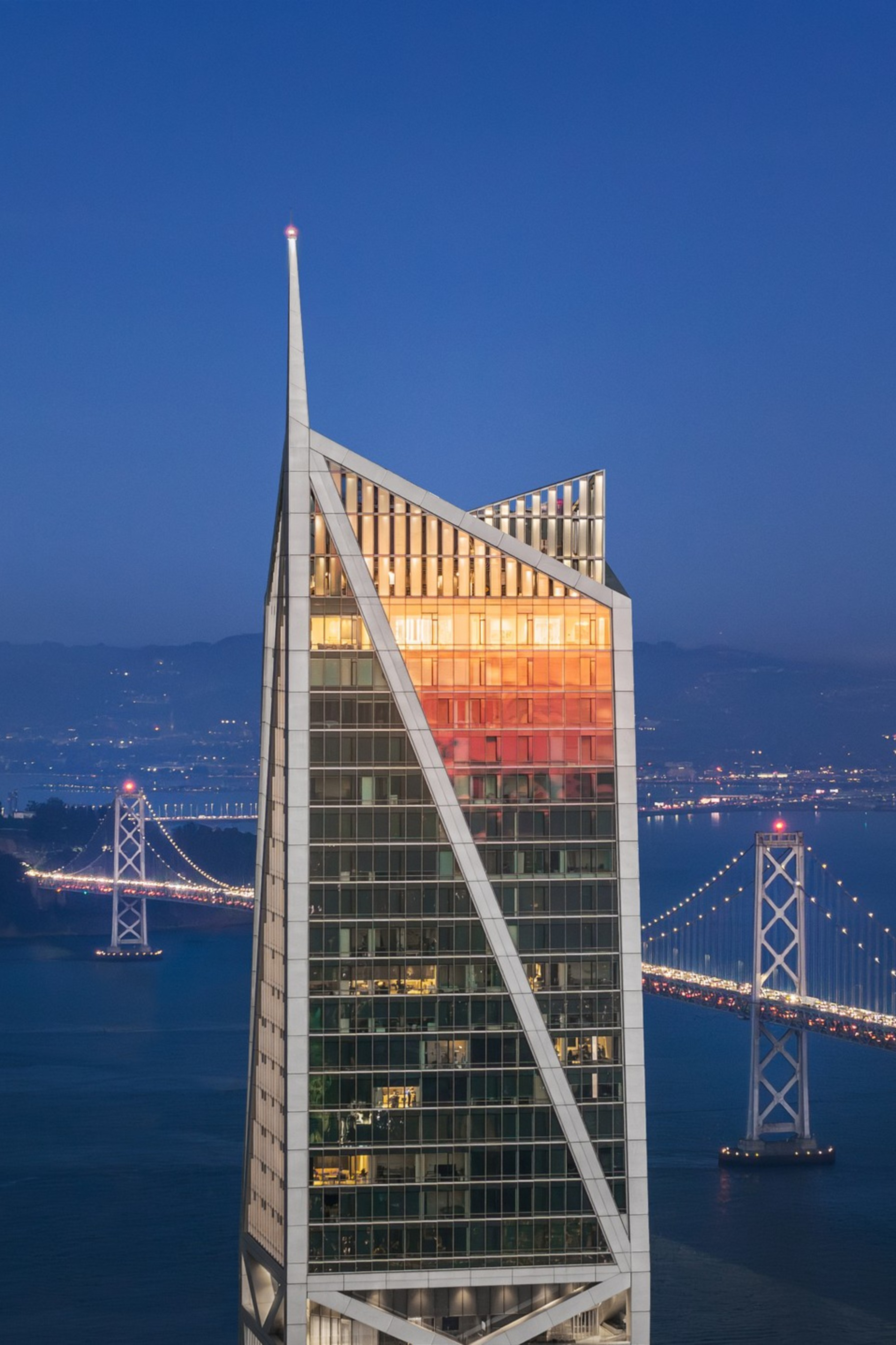 A tall, modern skyscraper reflects the sunset with a bridge and city lights in the background over a large body of water at dusk.