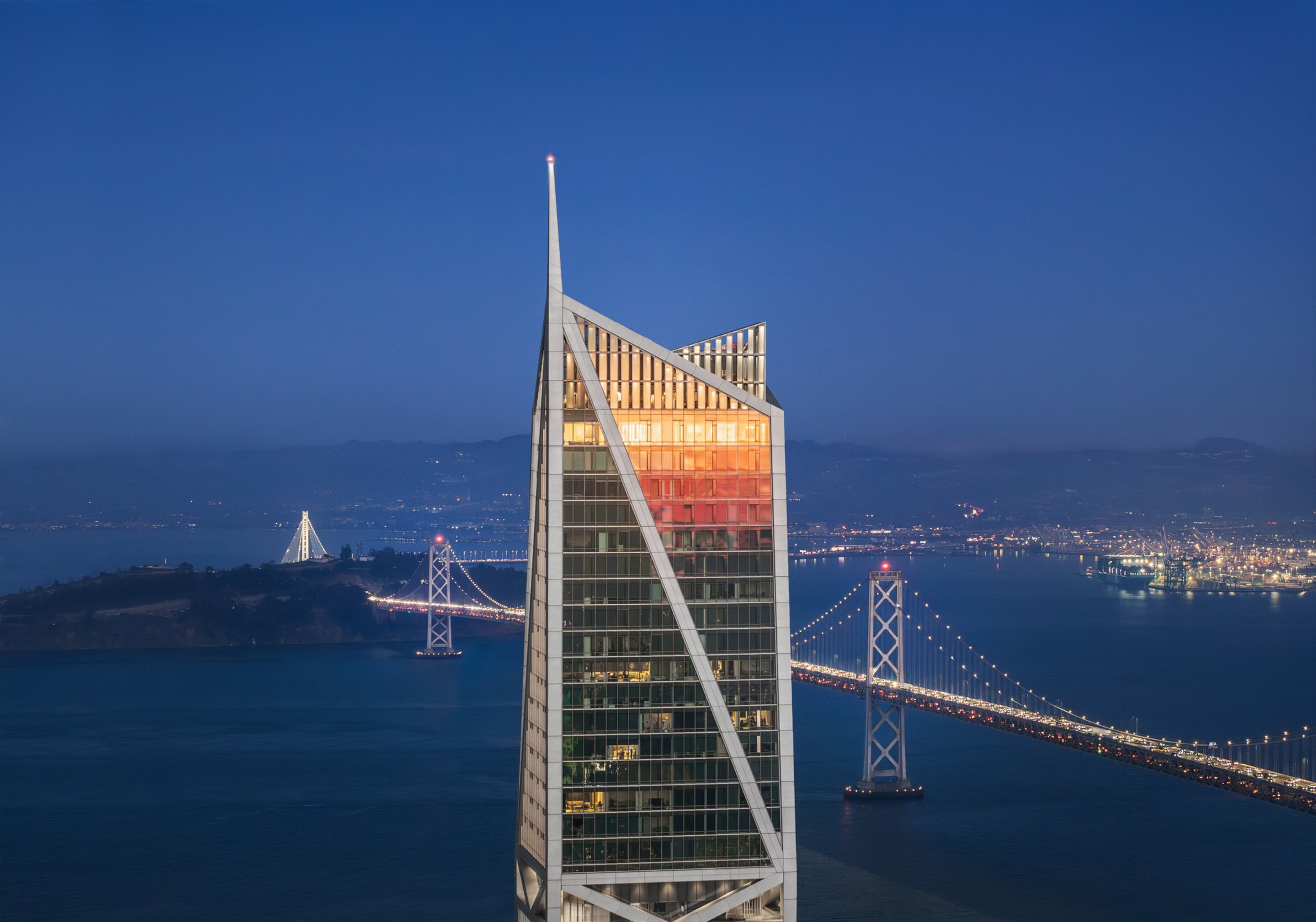 A tall, modern skyscraper reflects the sunset with a bridge and city lights in the background over a large body of water at dusk.