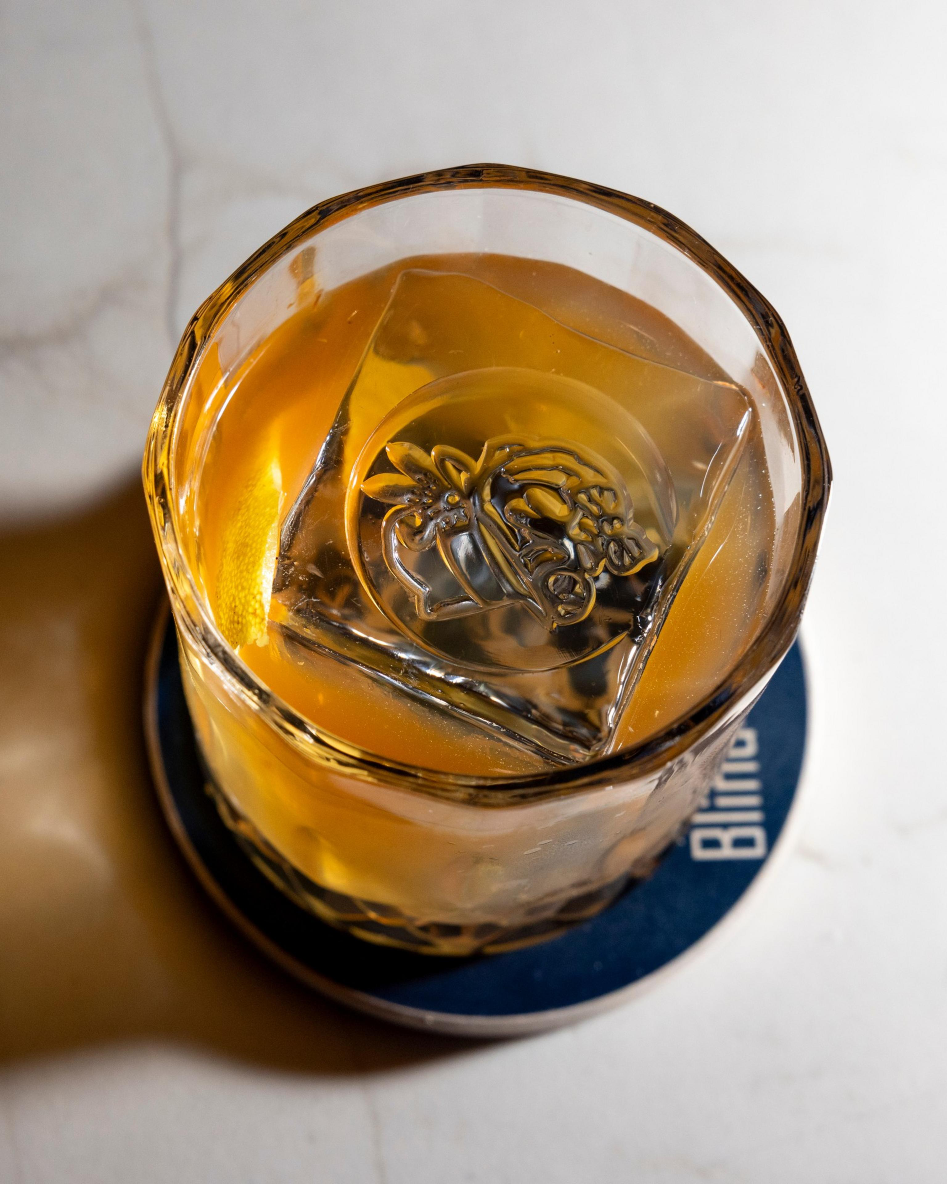A glass with an amber liquid, likely a cocktail, has a square ice cube featuring an intricate design. It rests on a round, blue coaster.