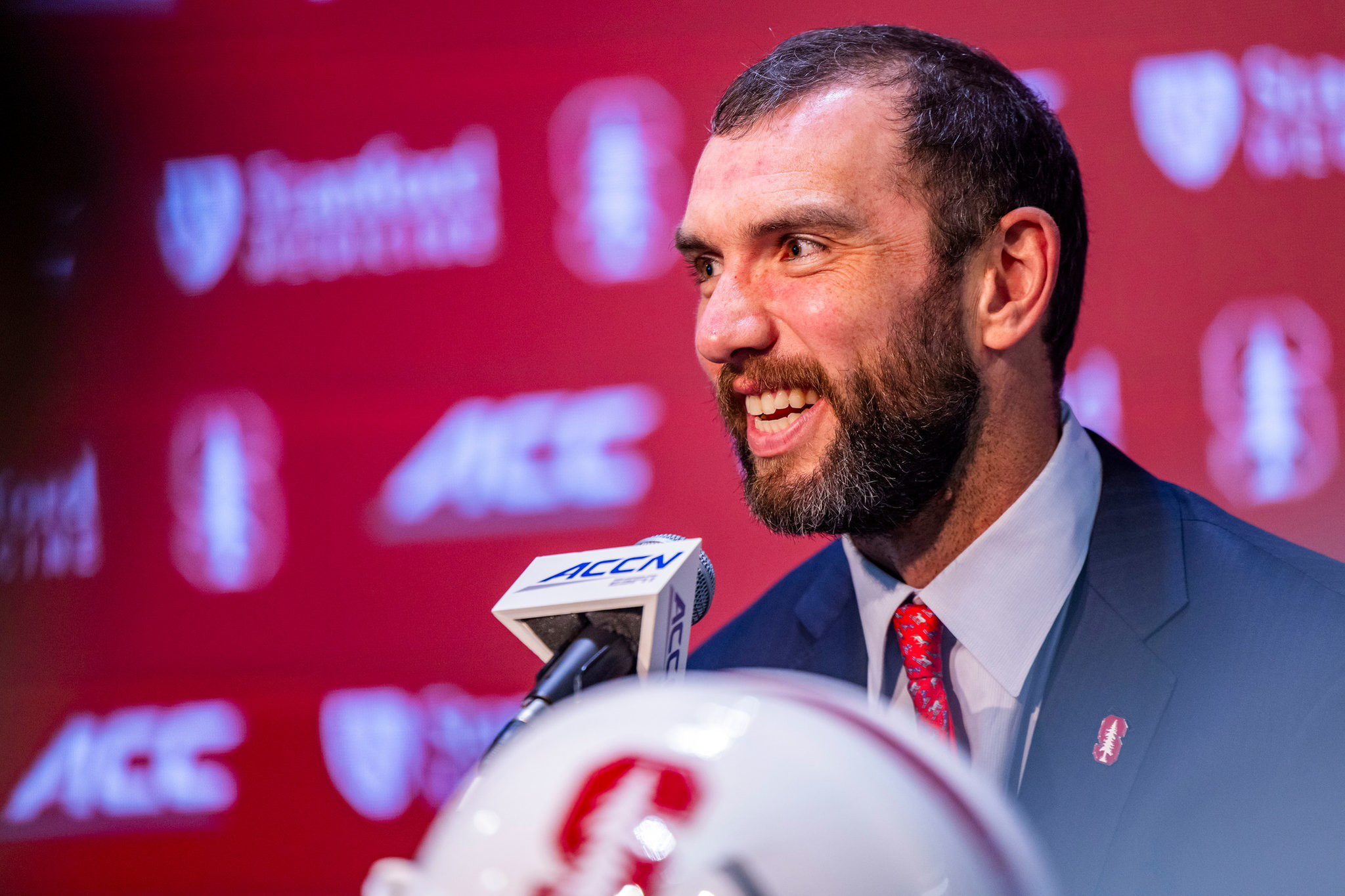 A man in a suit and red tie smiles while speaking into a microphone labeled “ACCN” with a white helmet and red background featuring Stanford logos.