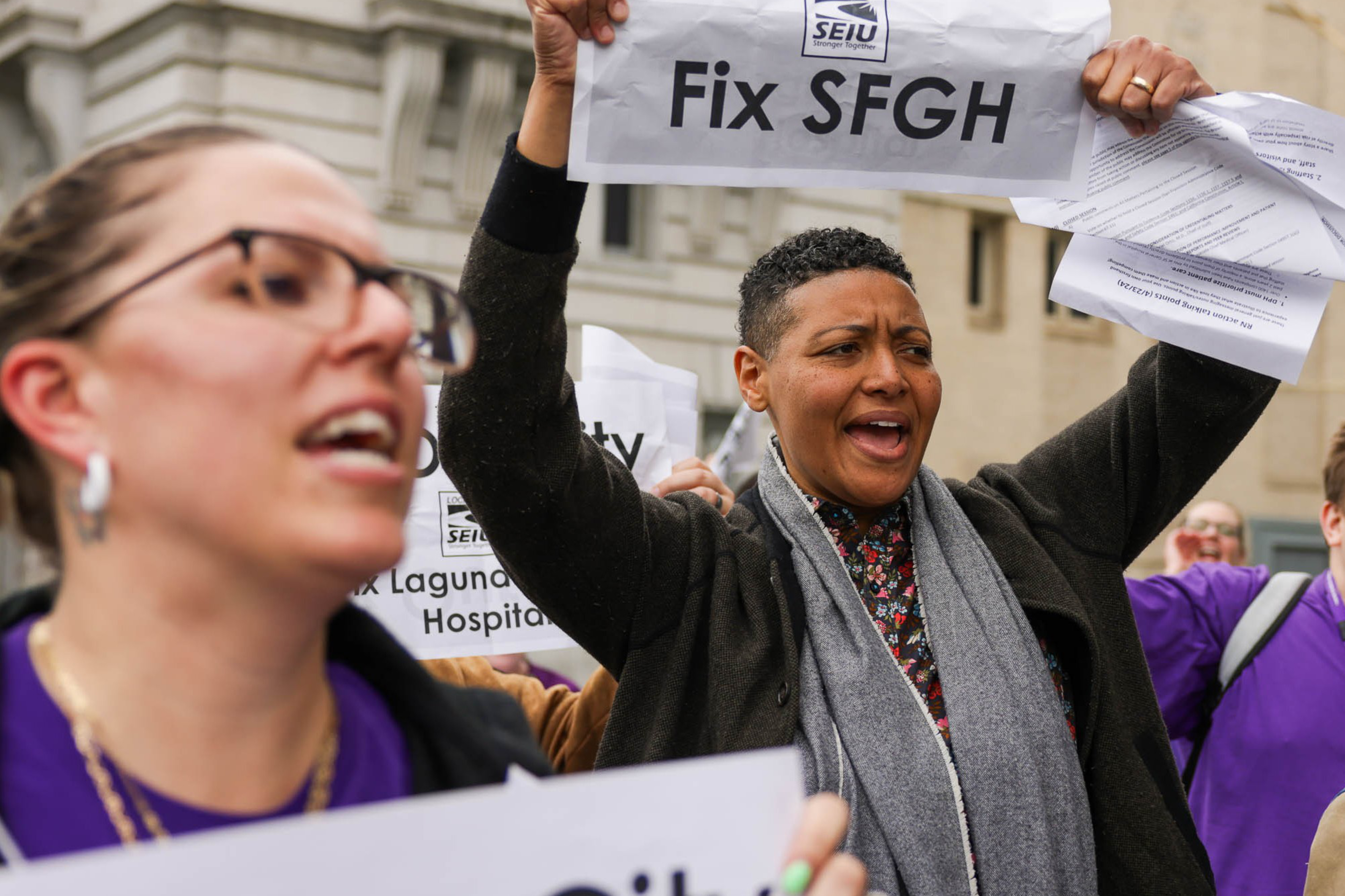 A group of people is protesting outdoors, holding signs reading "Fix Our City" and "Fix SFGH," with determined expressions and wearing purple clothing.