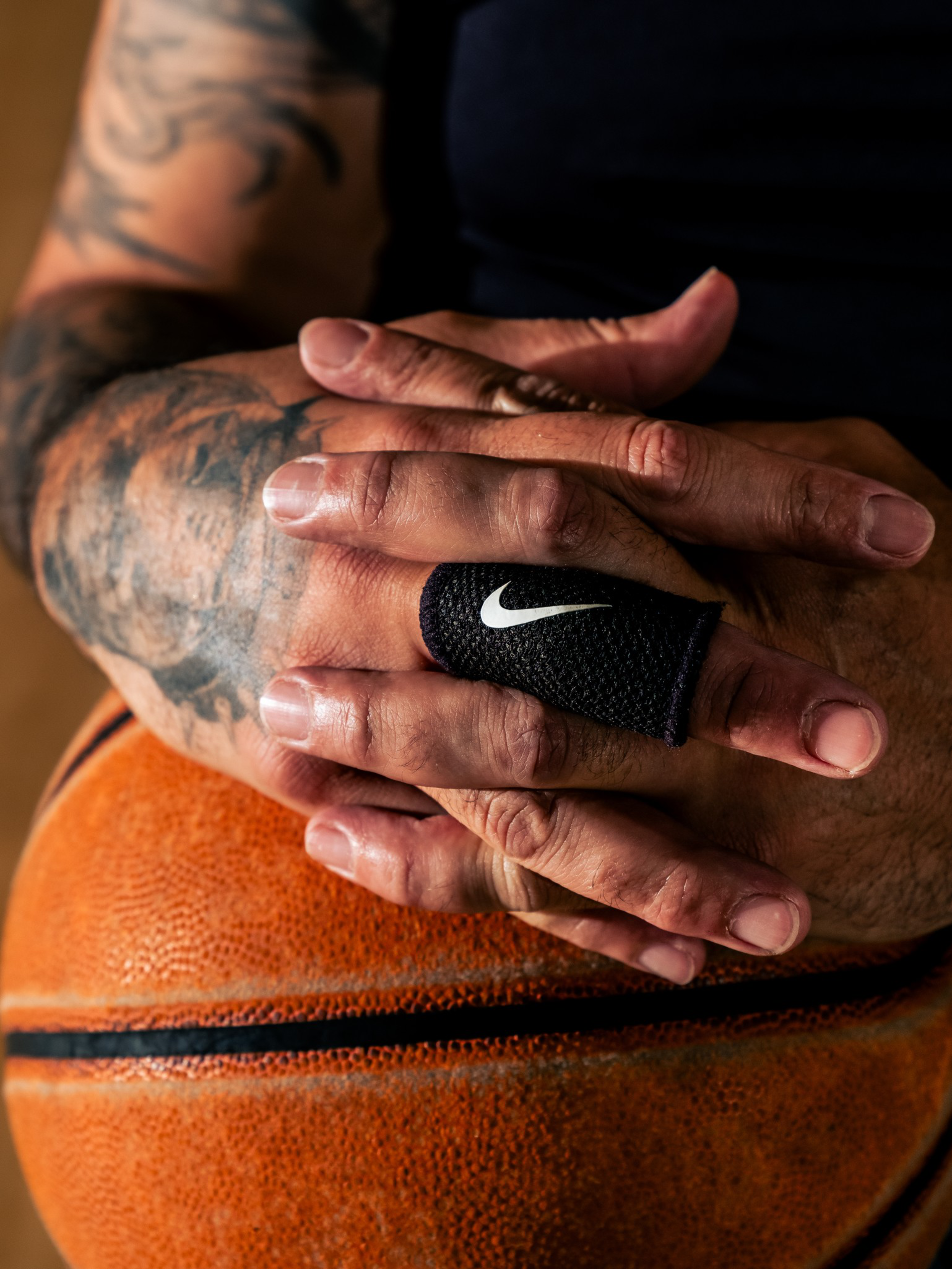 Tattooed hands resting on a basketball, with one finger wrapped in a black Nike-branded bandage, showing textured basketball surface.