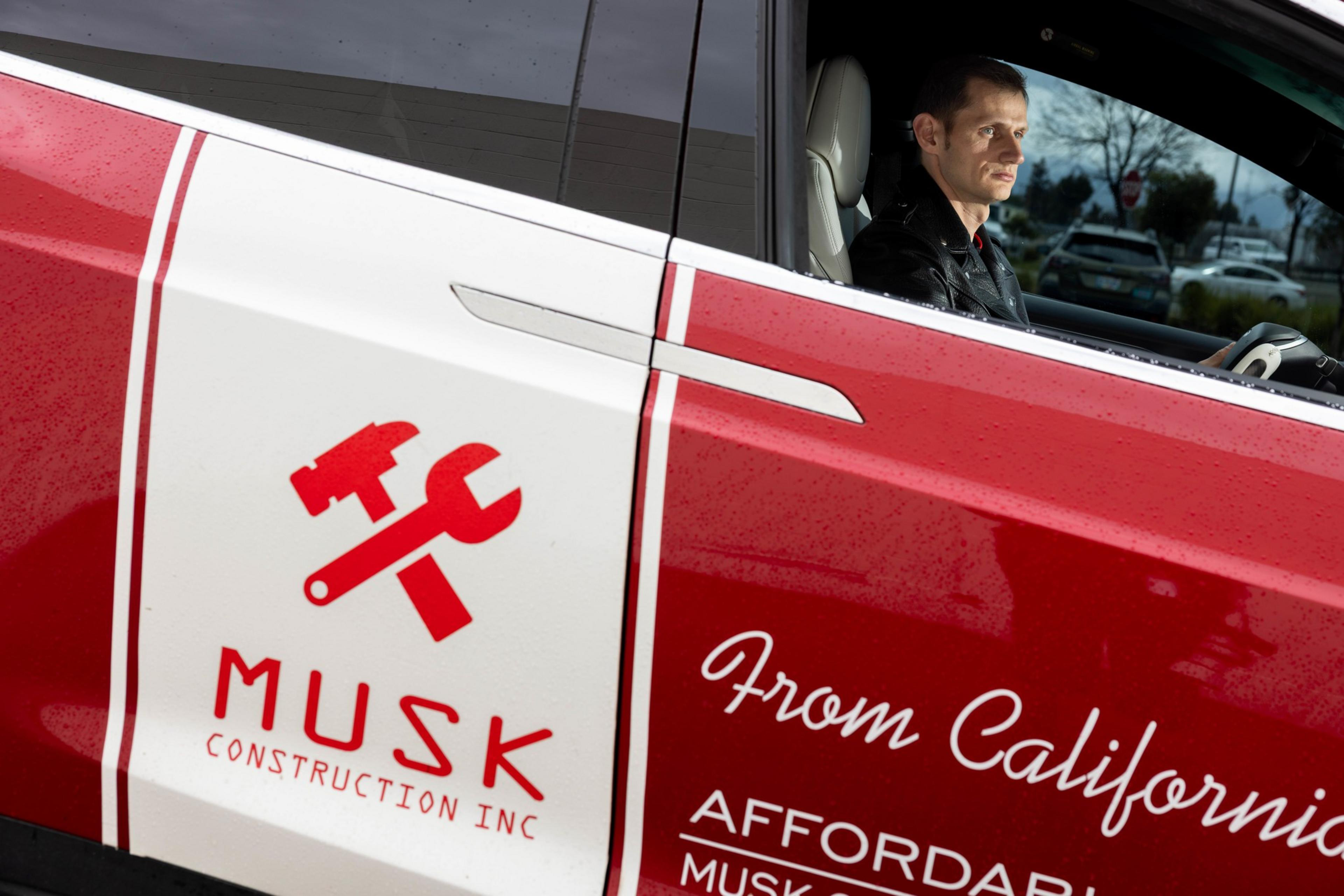 A man is sitting in a red vehicle with "Musk Construction Inc" branding, featuring hammer and wrench icons, parked in a lot.