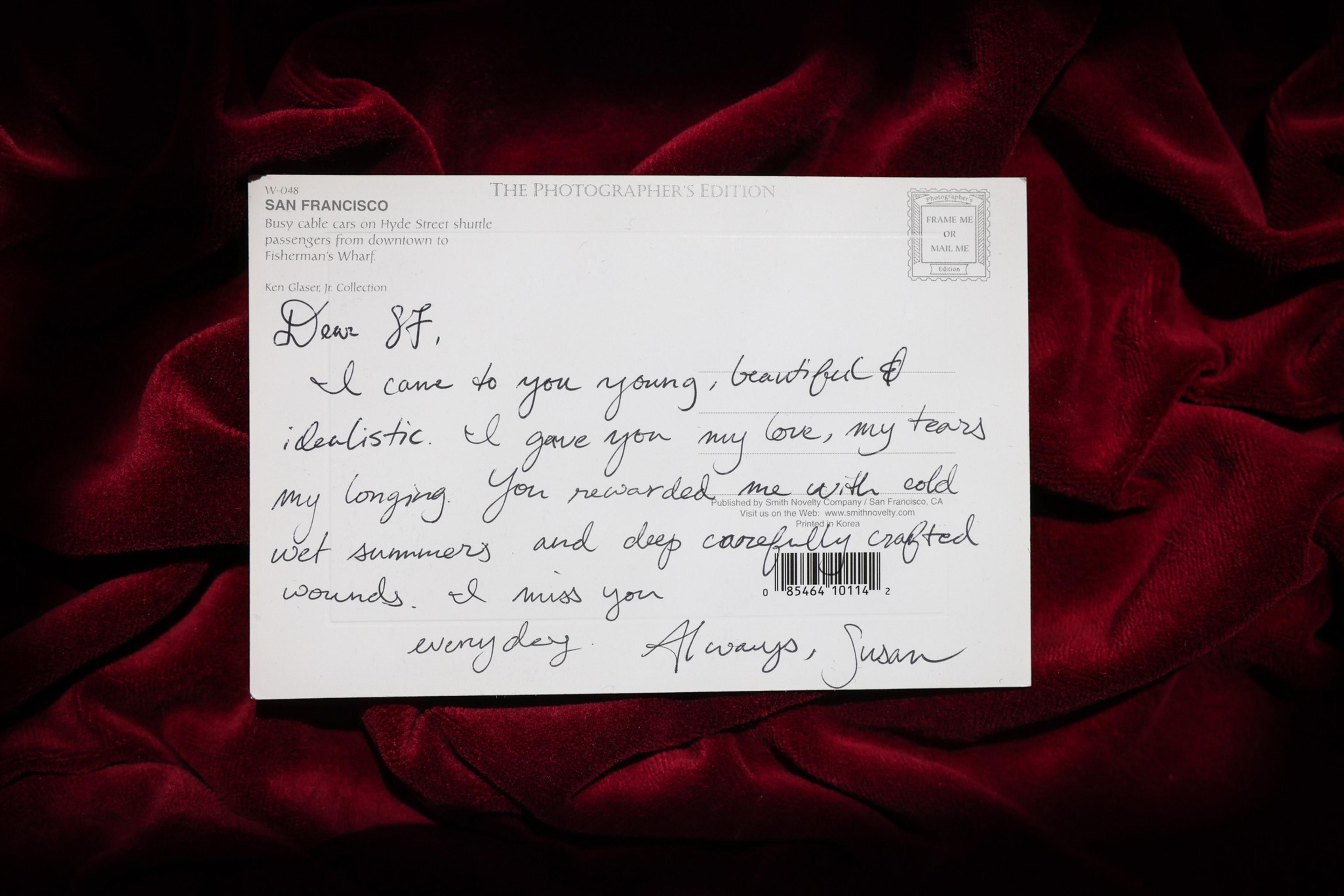A handwritten postcard on a red velvet fabric reads, expressing love, longing, and missing San Francisco with mentions of cold summers and deep wounds.