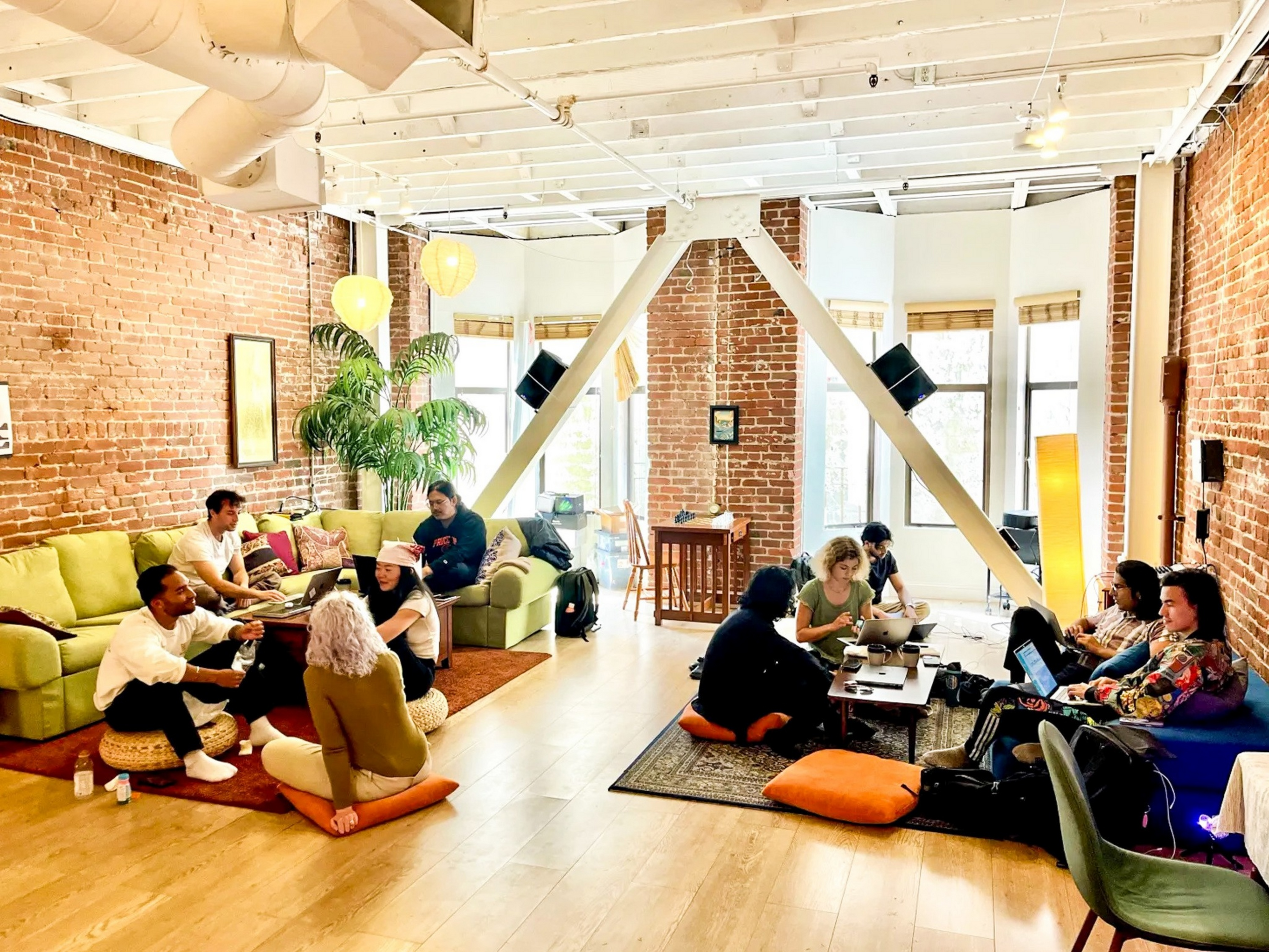 People work and chat in a bright, spacious room with exposed brick walls, green couches, cushions on the floor, plants, and laptops on low tables.