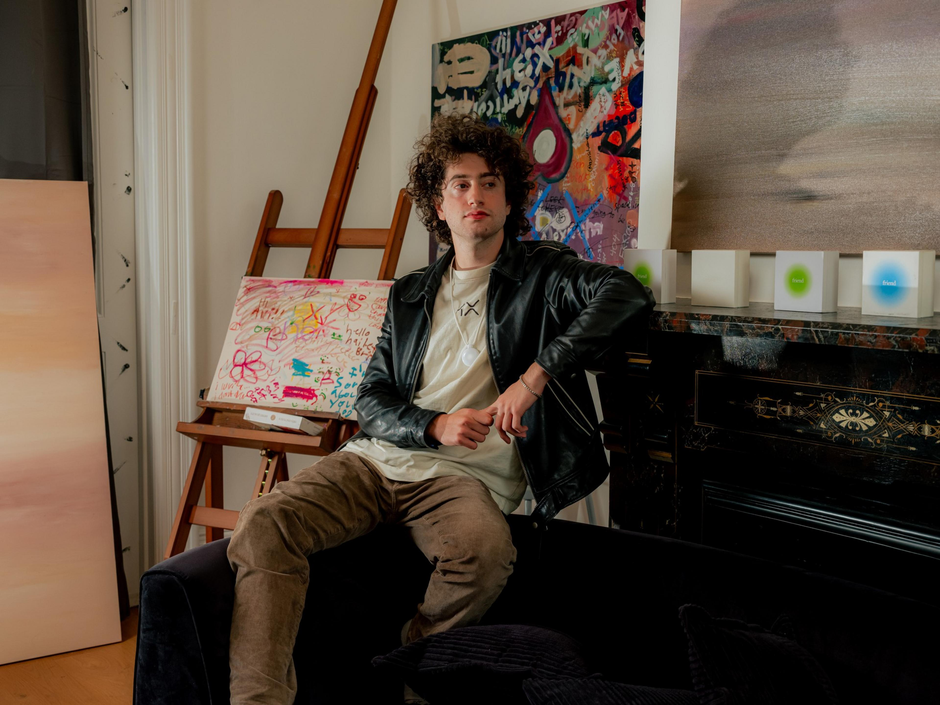 A young man with curly hair wears a black leather jacket and beige pants, sitting on a couch near colorful abstract paintings on easels.