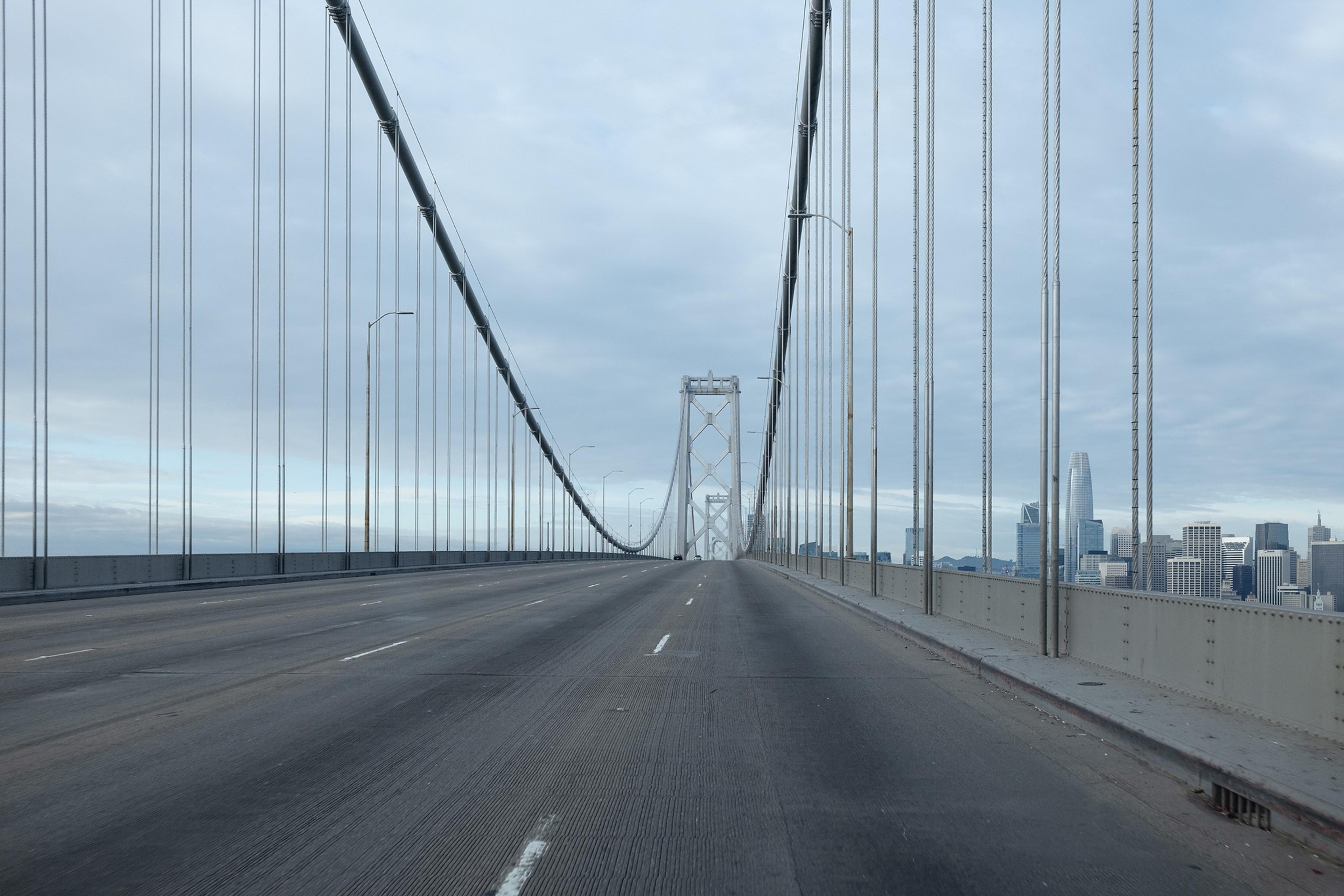 A wide view of an empty suspension bridge with vertical cables extending upward, leading towards a city skyline under a cloudy sky.
