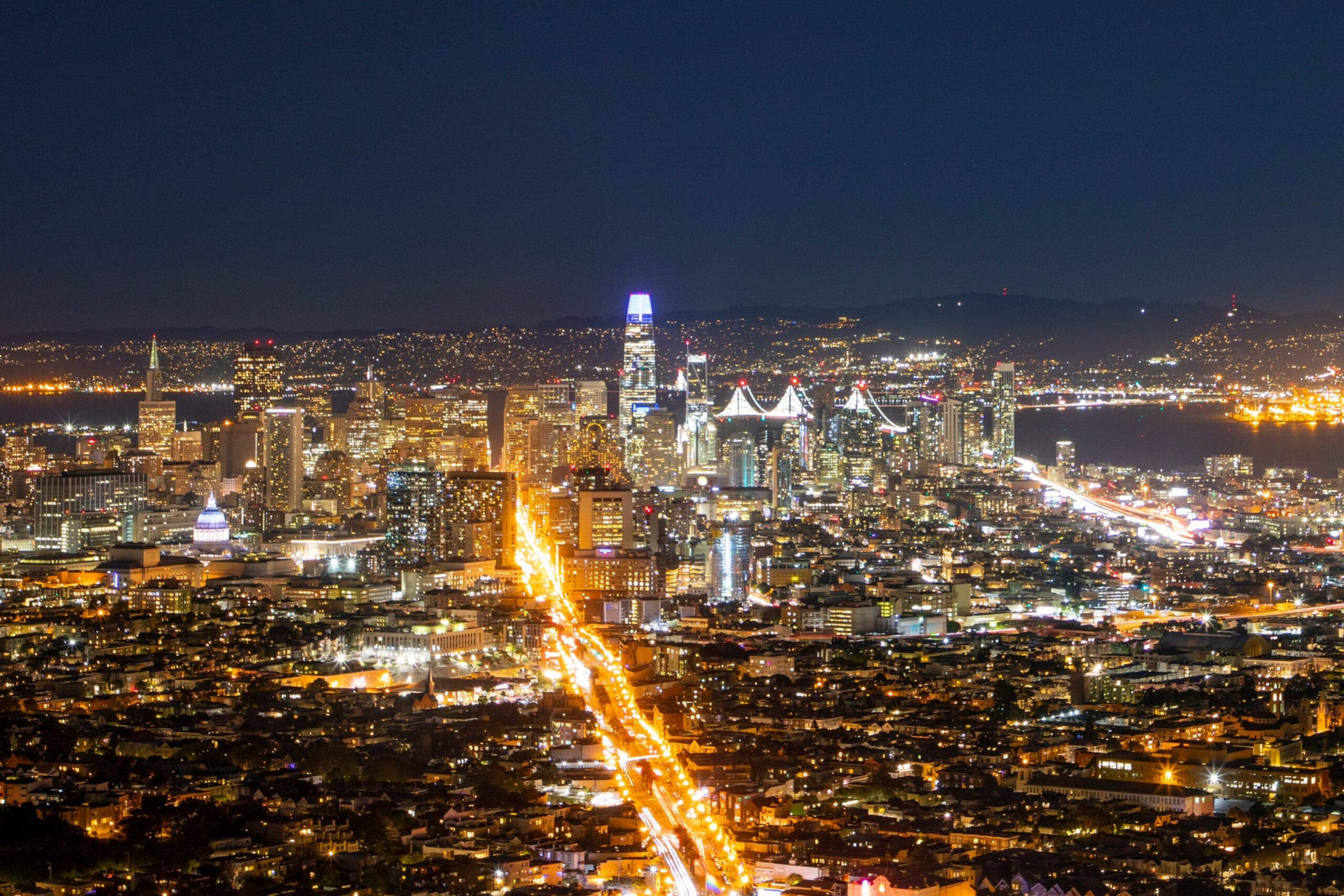 A night view of San Francisco with bright lights, towering skyscrapers, a lit-up bridge in the distance, and busy traffic arteries.