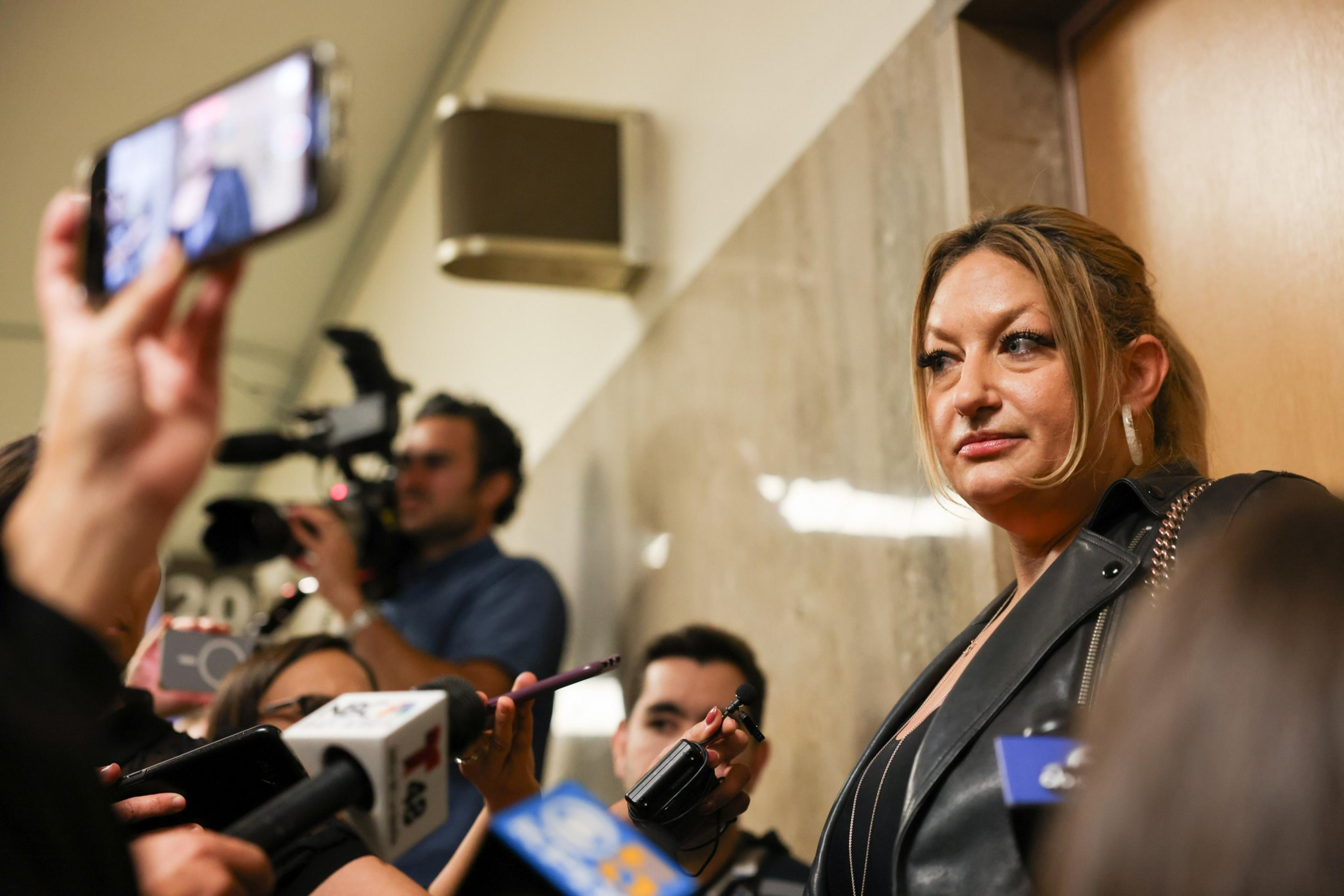 A woman in a black leather jacket stands surrounded by reporters and microphones. A camera crew and people with phones capture the scene, creating a busy atmosphere.