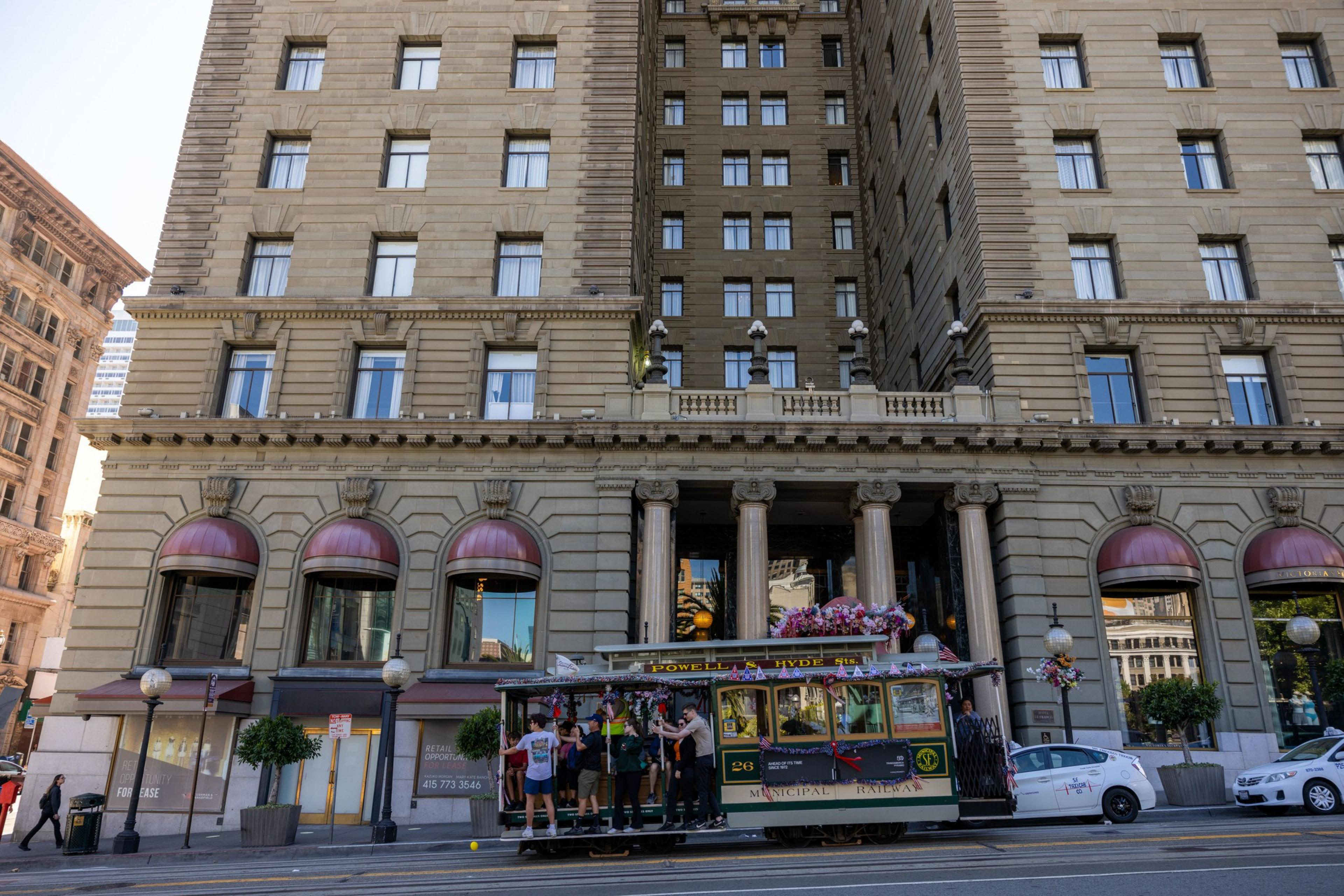 A decorated, vintage trolley car filled with people is passing by a grand, historic building with large windows and red awnings on a busy city street.