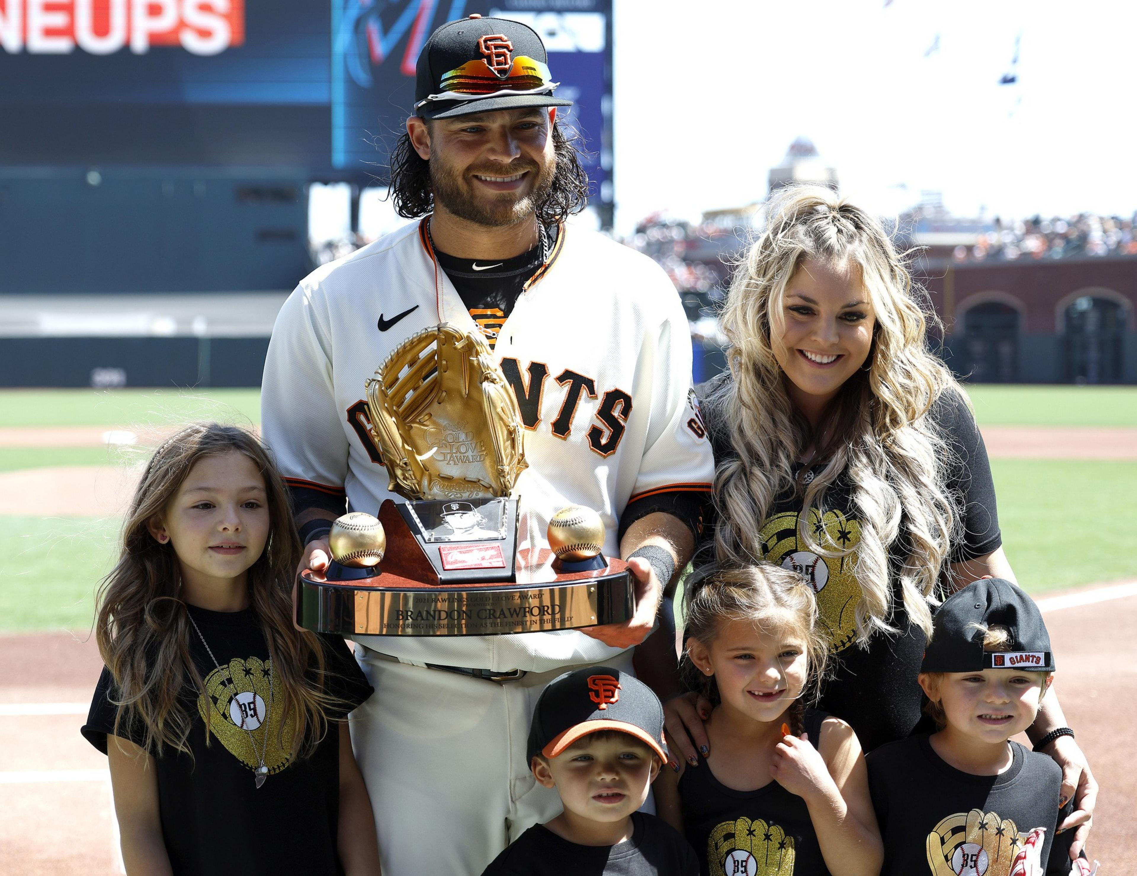 A baseball player in a Giants uniform holds a gold glove trophy. He is surrounded by a smiling woman and four children wearing matching baseball-themed shirts.
