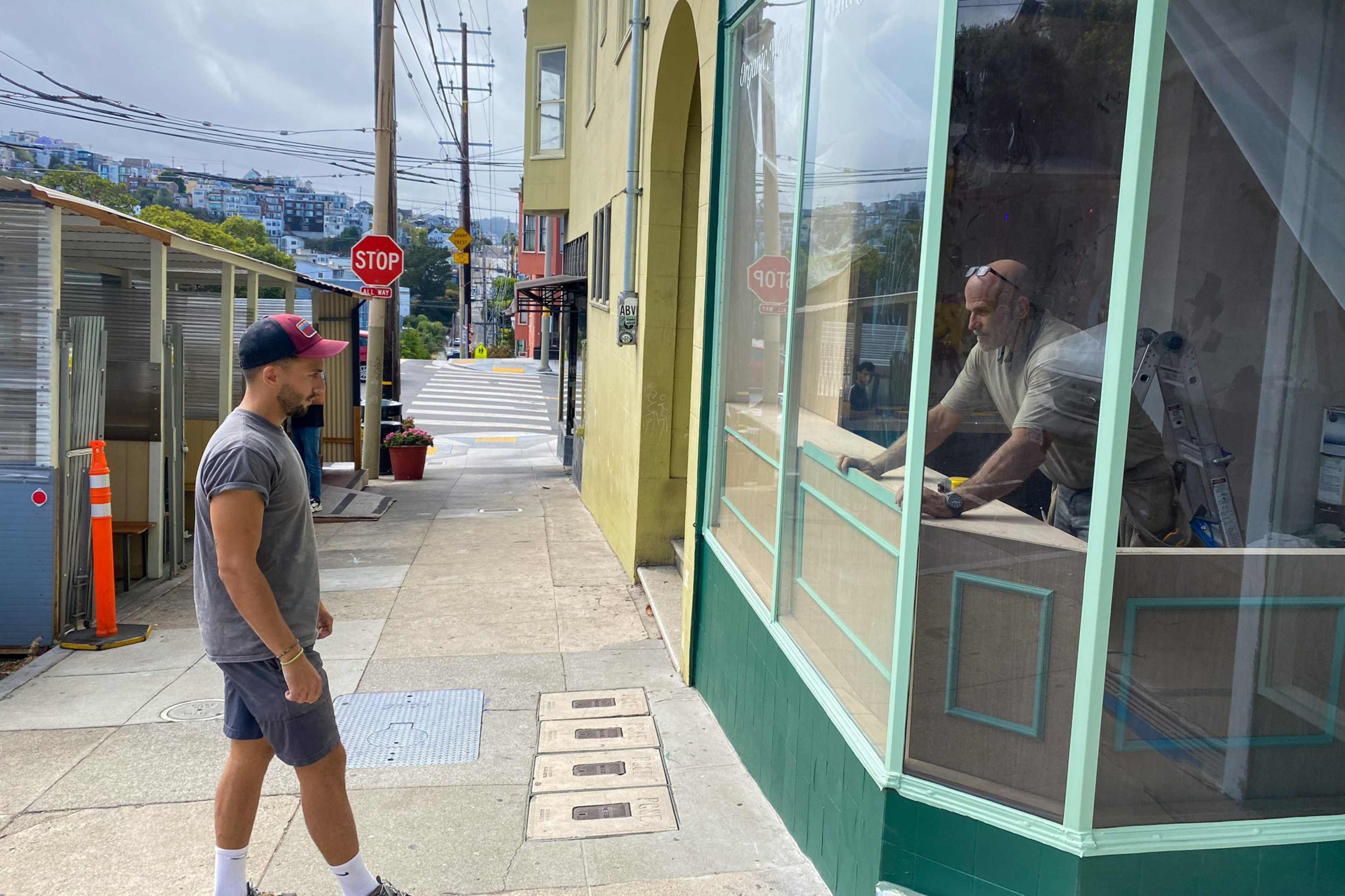 A man in a gray shirt and shorts stands on a sidewalk looking into a large window where another man is working inside a building.