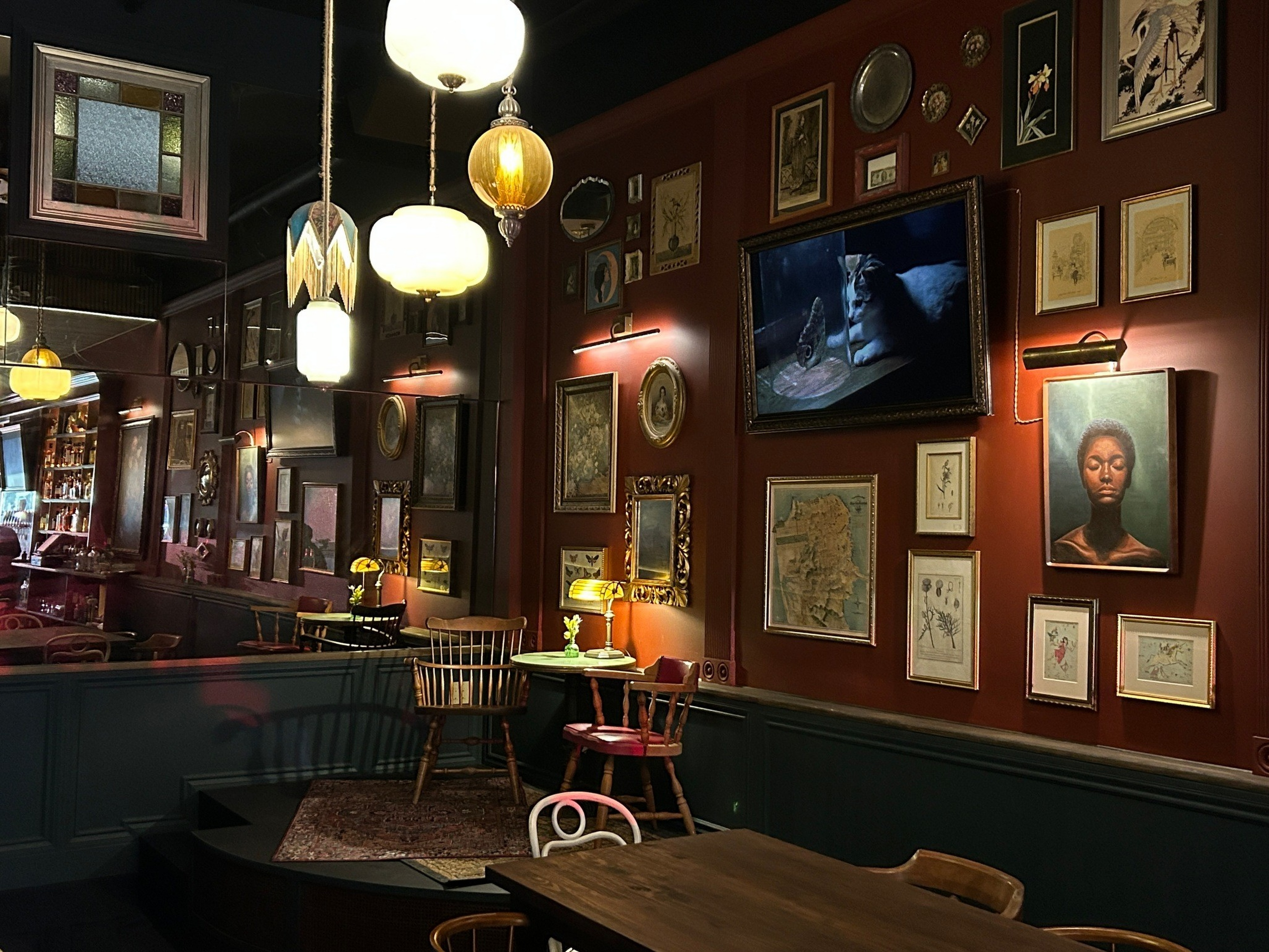 A cozy room with dim lighting features a wall covered in eclectic framed artwork, wooden chairs around small tables, and vintage hanging lamps.