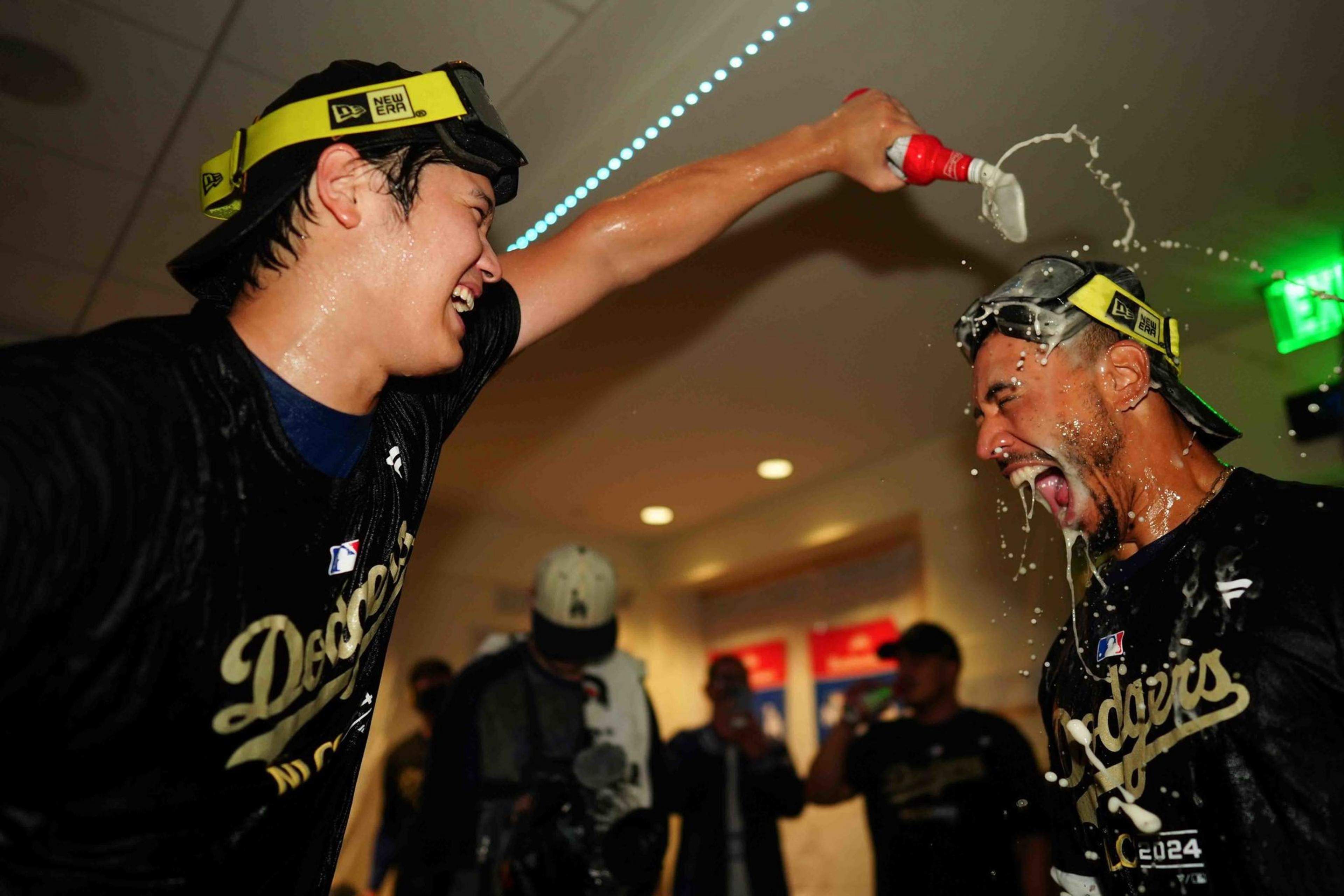 Two men celebrate in a room; one pours a drink over the otherâ€™s head. Both wear goggles and black shirts with "Dodgers" written on them.