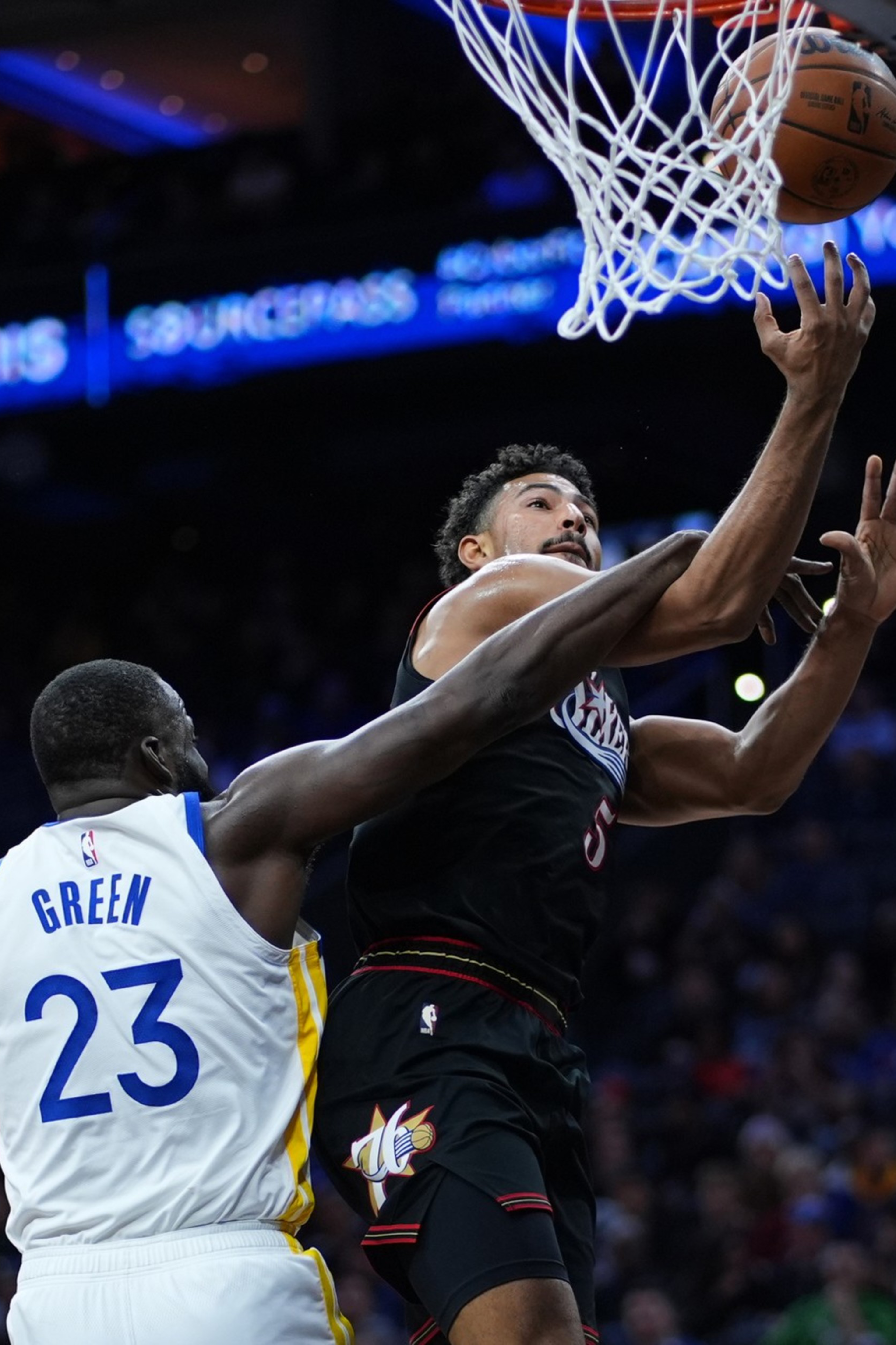 A basketball player in a black jersey leaps to score as a defender in a white jersey with "Green 23" reaches to block him near the hoop.