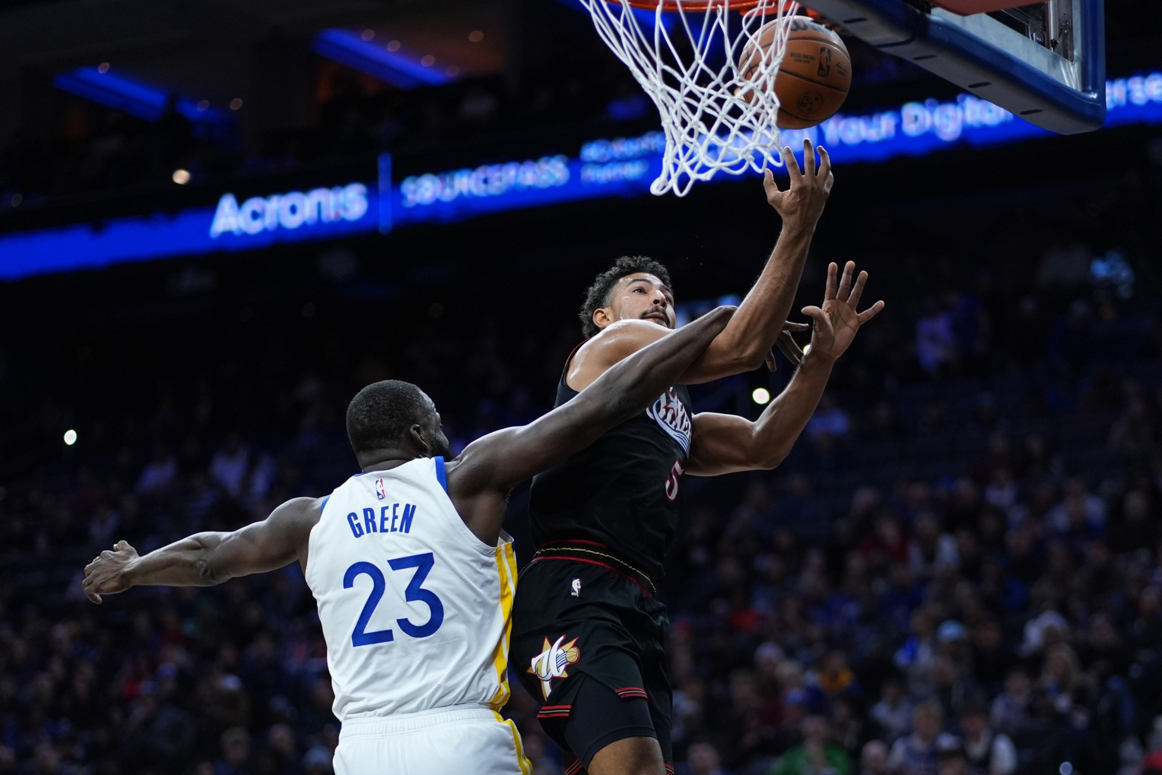 A basketball player in a black jersey leaps to score as a defender in a white jersey with "Green 23" reaches to block him near the hoop.