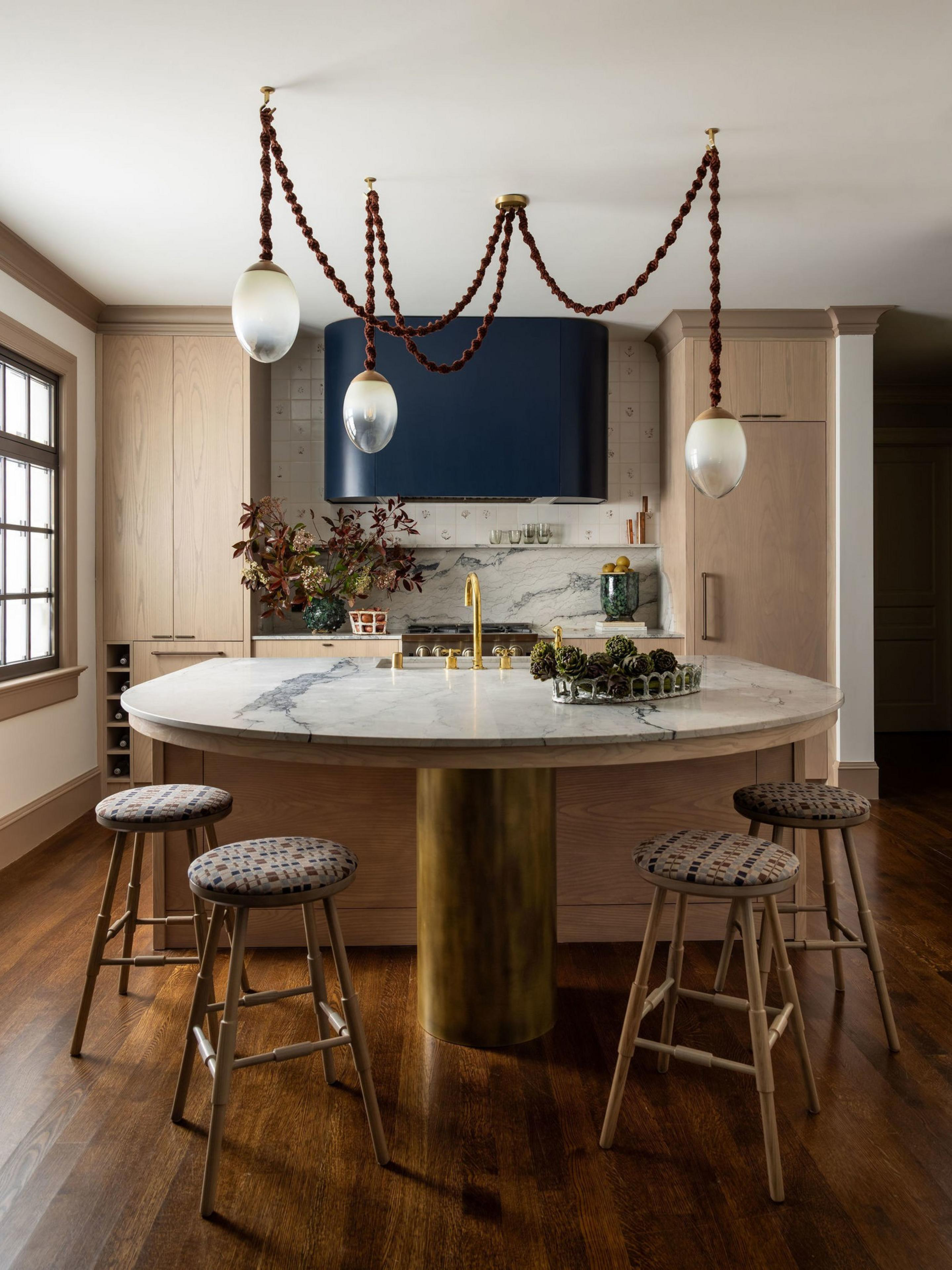 A modern kitchen features a round marble island with a brass pedestal, four patterned stools, light wood cabinets, a blue hood, and twisted rope pendant lights.