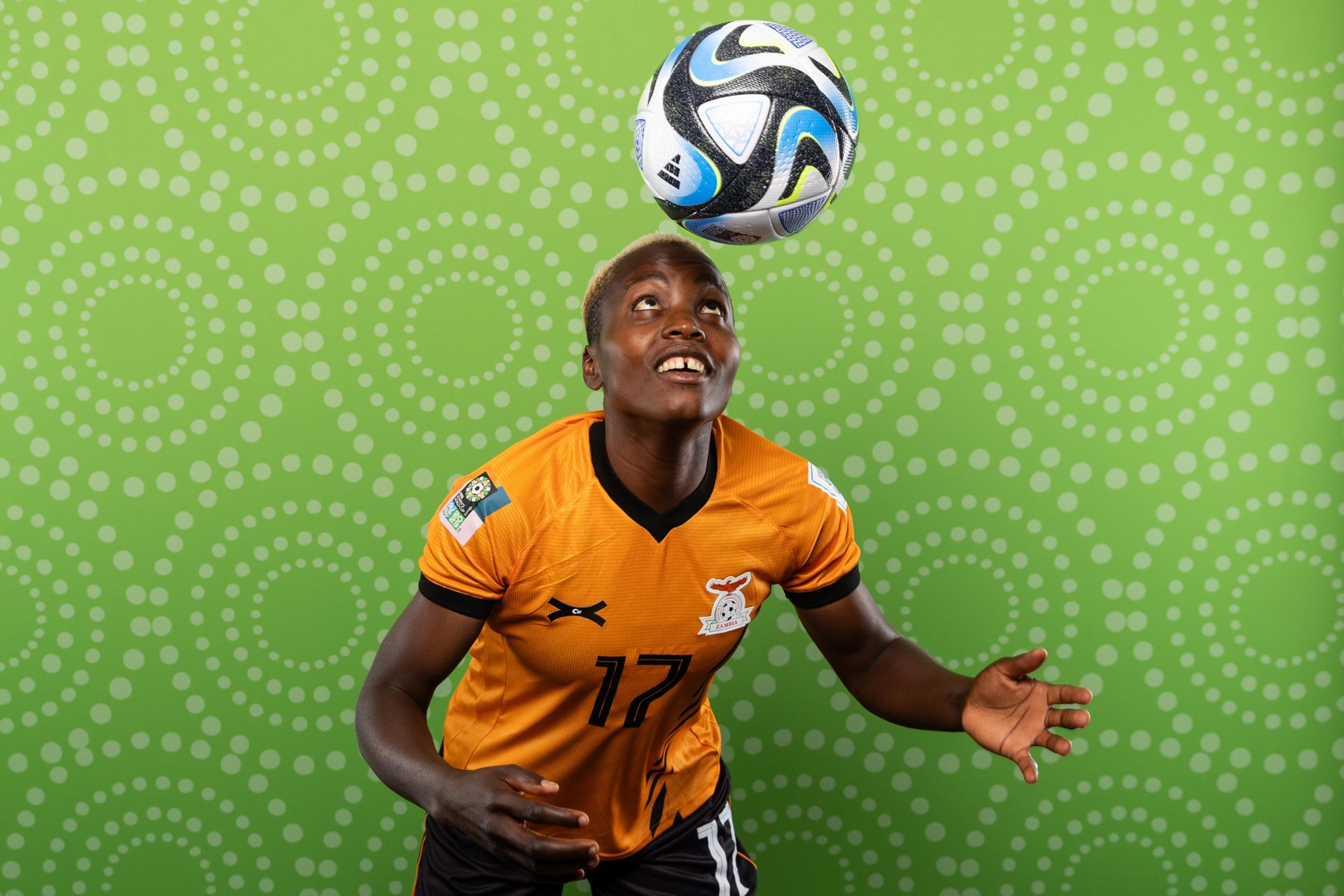 A soccer player in an orange jersey with number 17 balances a black, white, and blue ball on their forehead against a green patterned background.