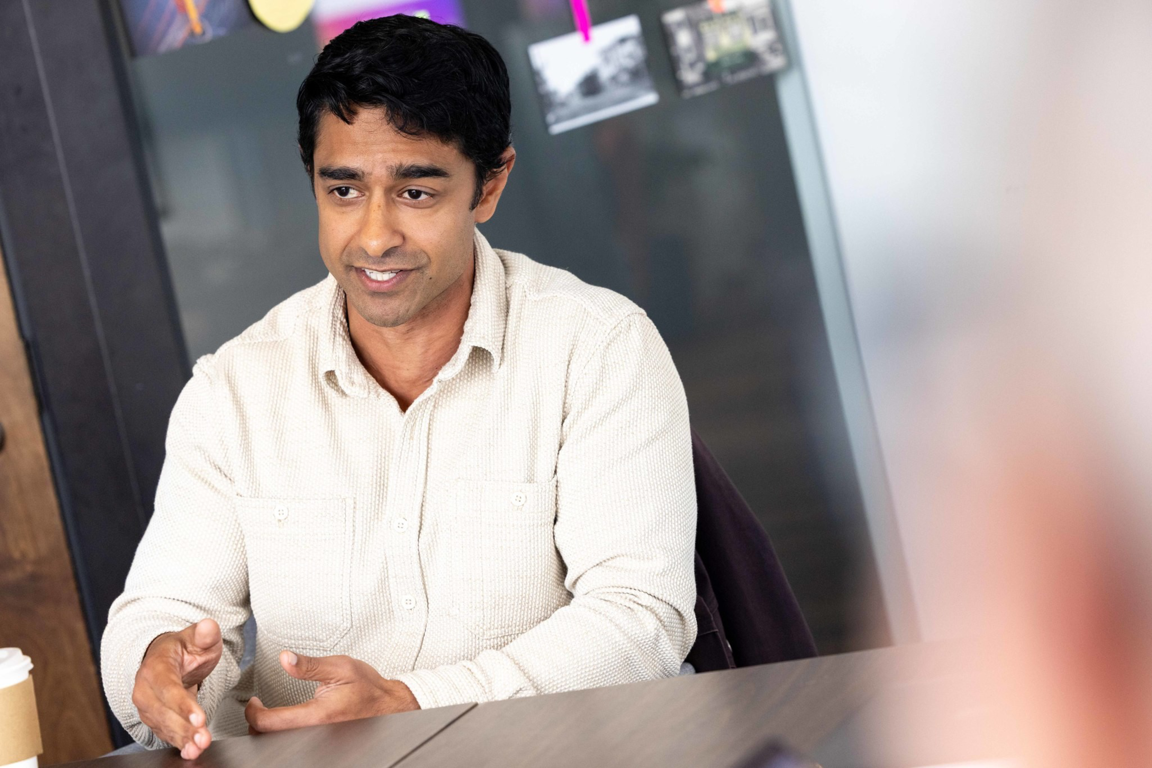 A man in a beige shirt gestures with his hands while speaking at a table, with photos clipped on a string in the blurred background.