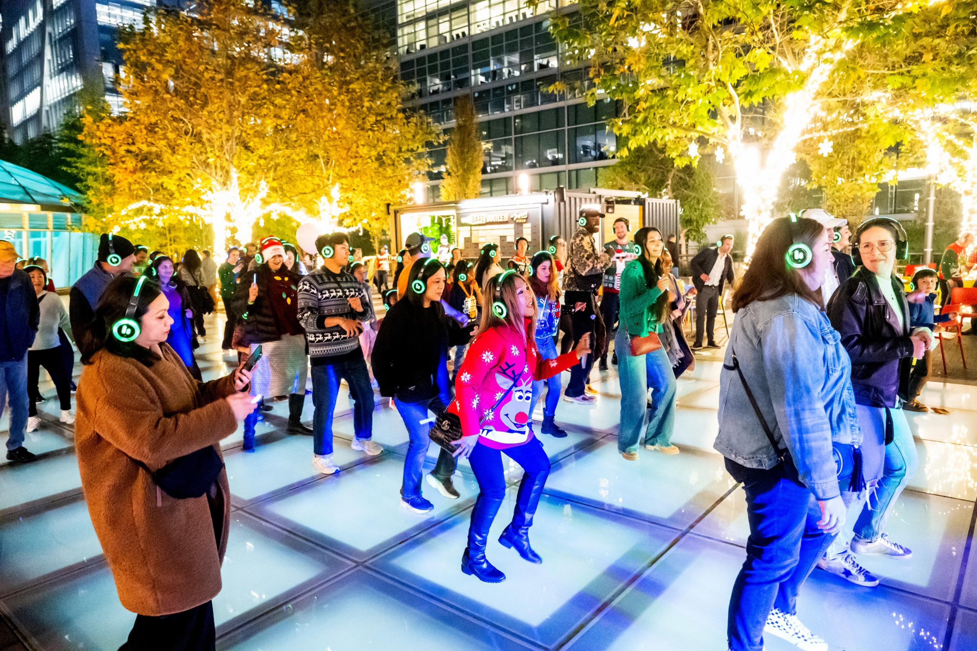 People are dancing at a festive outdoor silent disco wearing glowing headphones. There's colorful lighting, holiday sweaters, and a backdrop of illuminated trees.